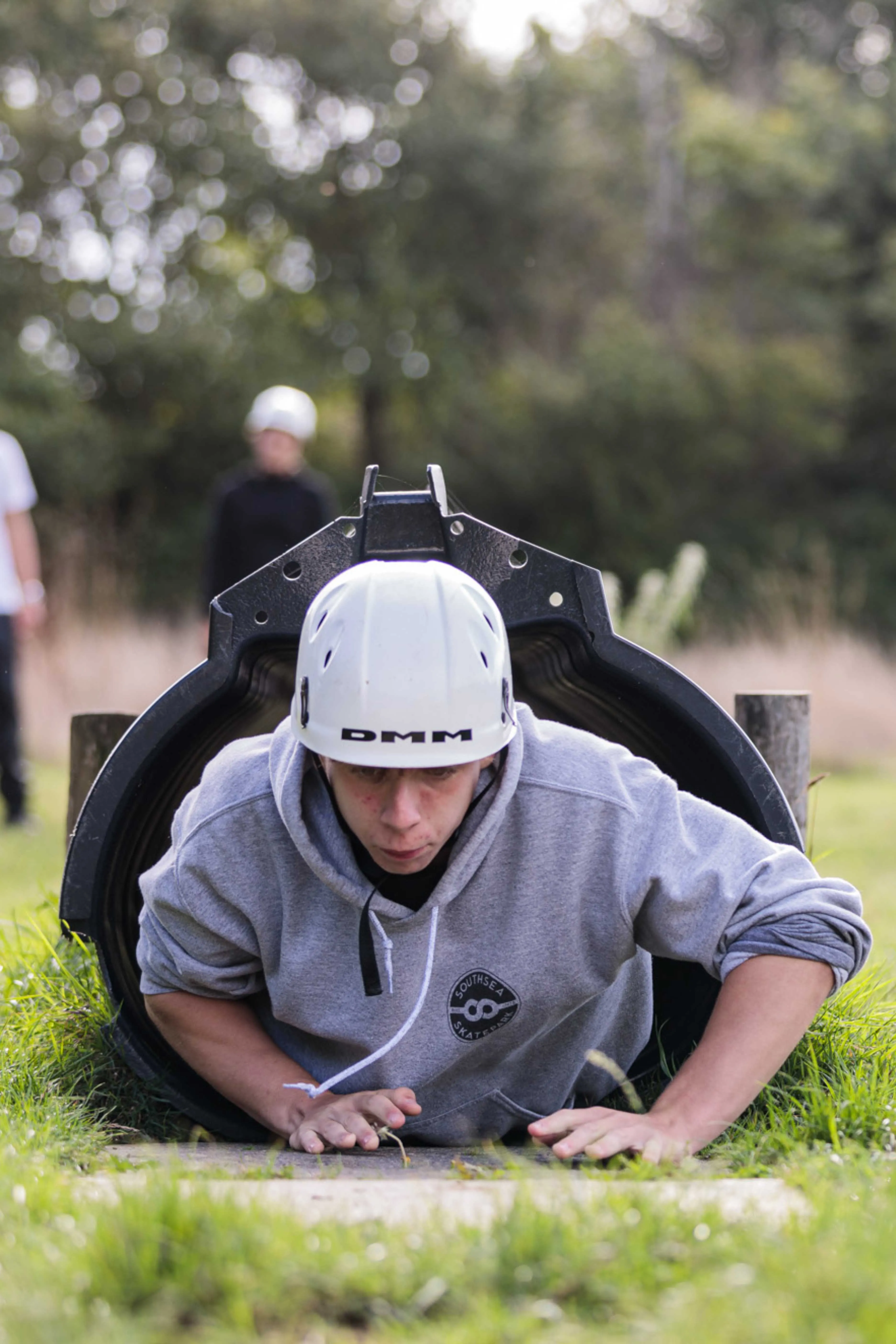 Person wearing a white helmet and grey hoodie crawling out of a black tunnel on green grass during an outdoor activity.