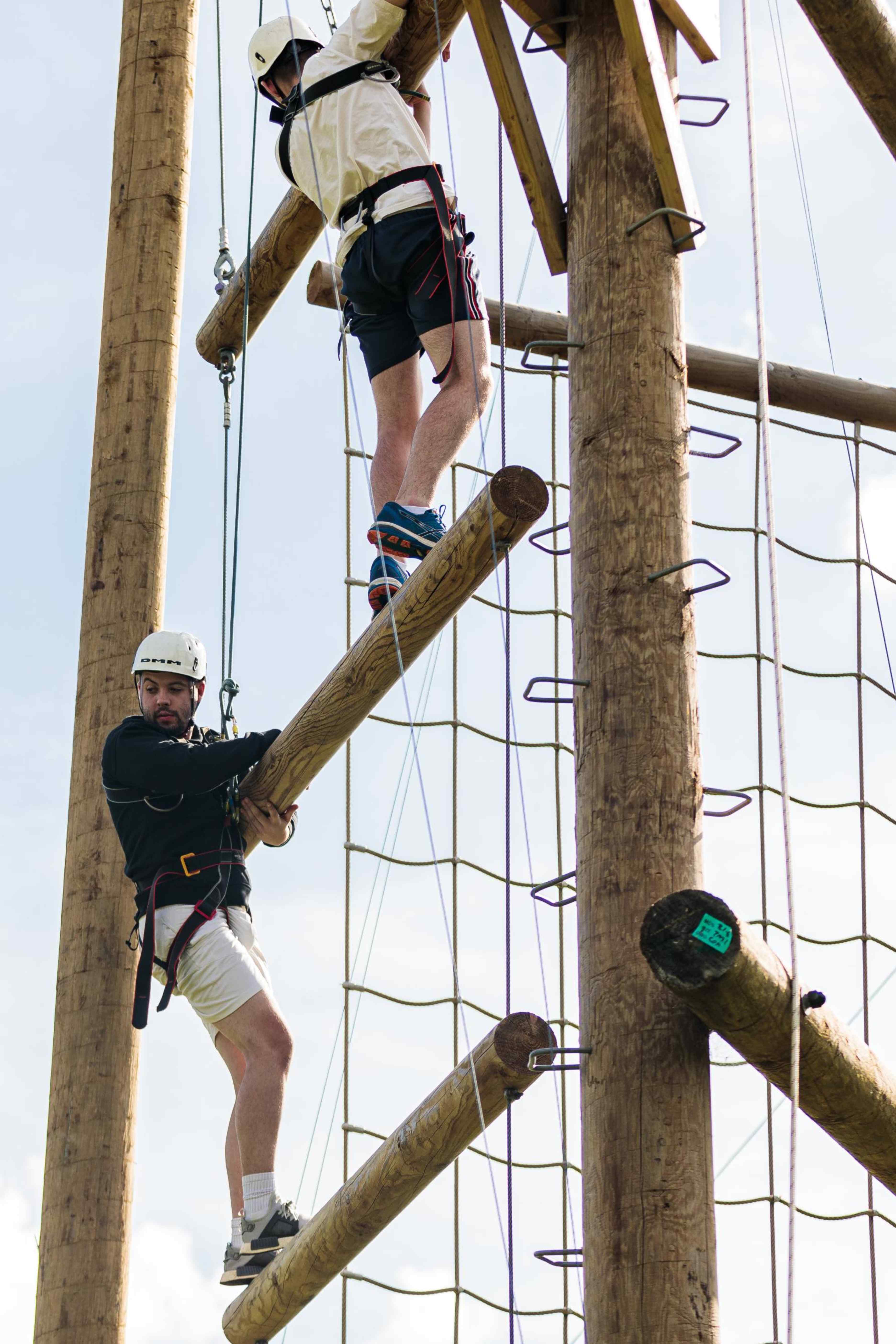 Two people wearing helmets and harnesses climbing wooden poles on a ropes course outdoors.