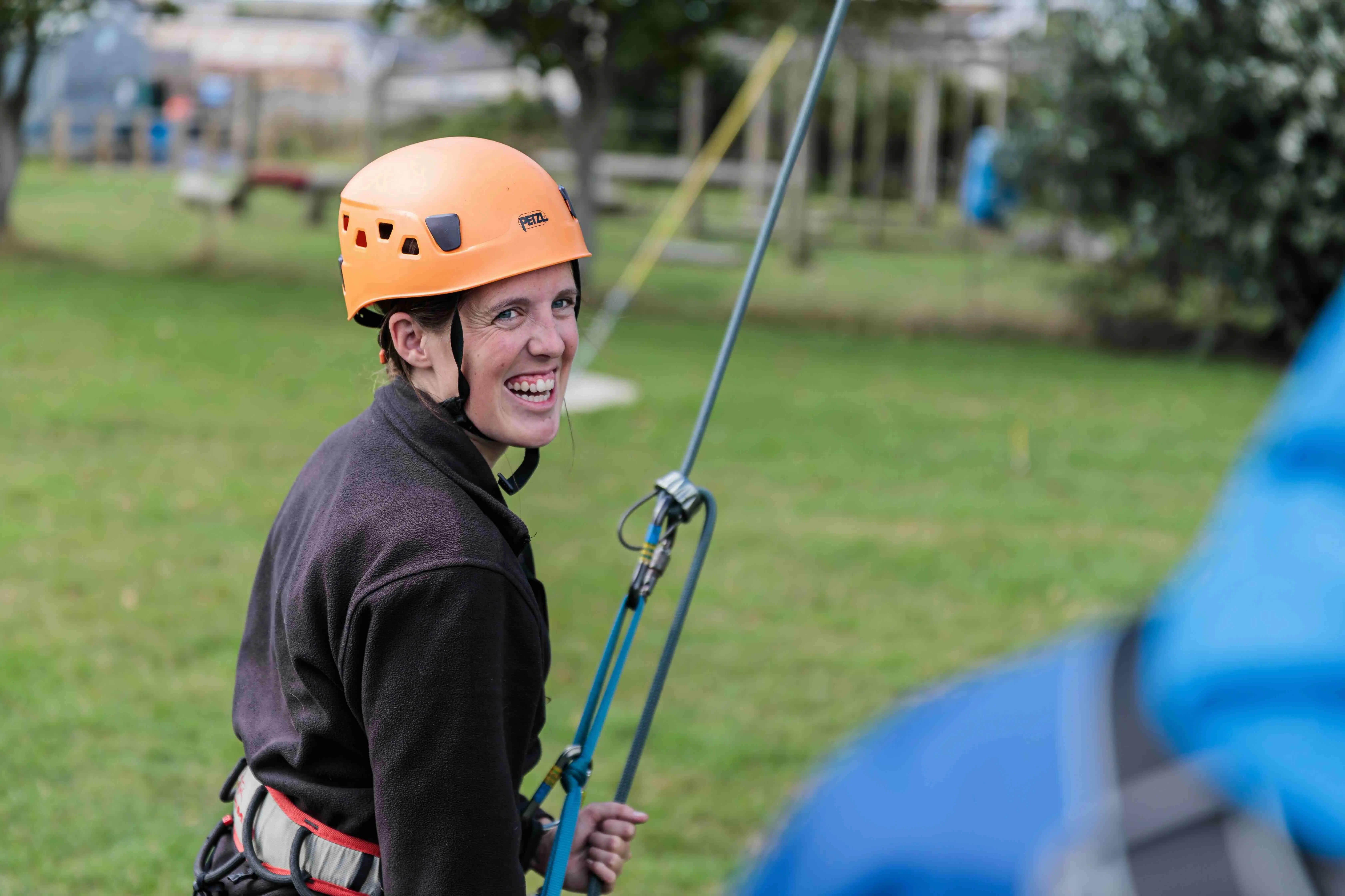 Smiling woman wearing an orange climbing helmet and harness holding a rope outdoors.