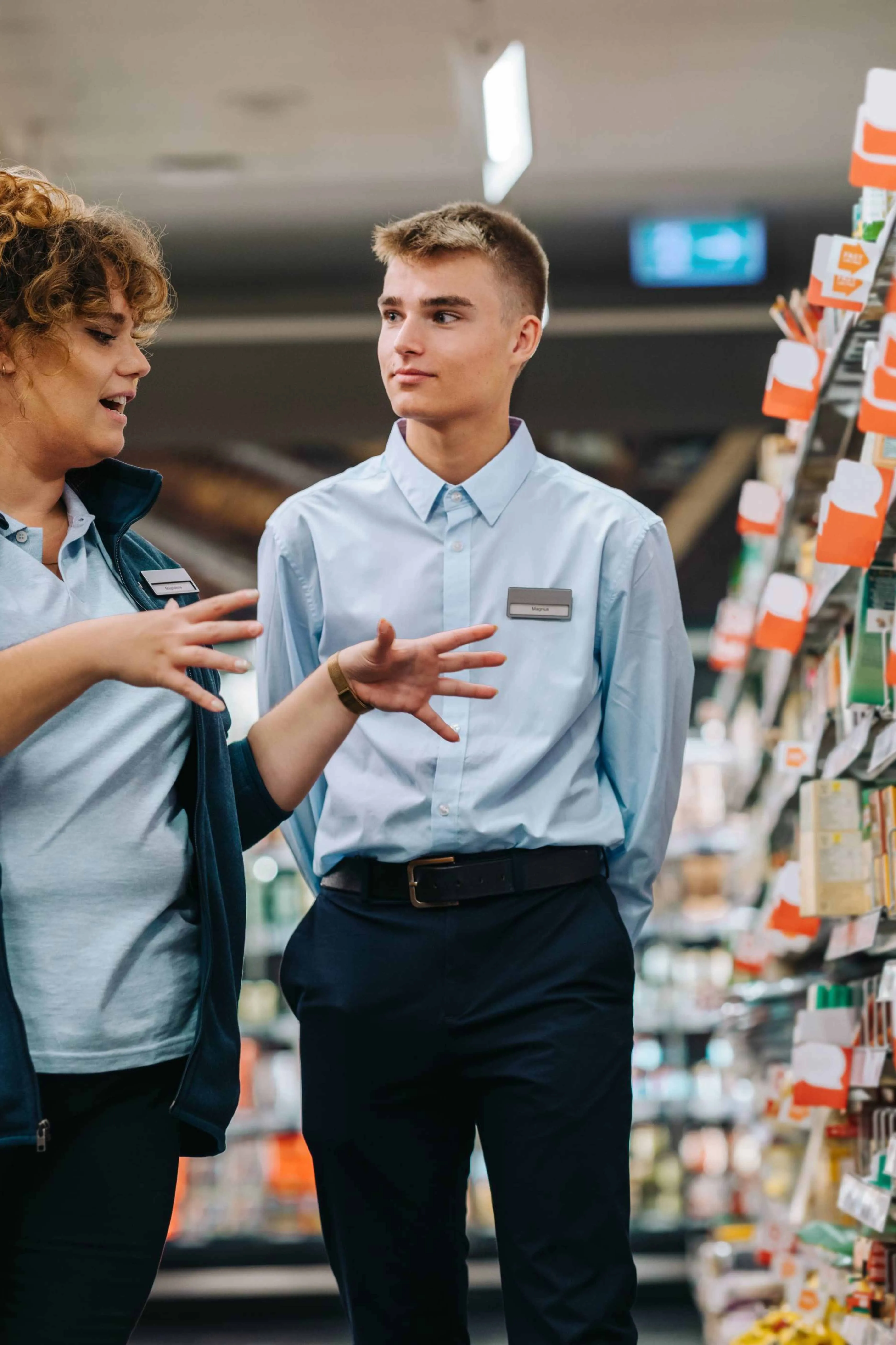 Two employees in light blue uniforms with name tags discussing in a store aisle with shelves stocked with products.