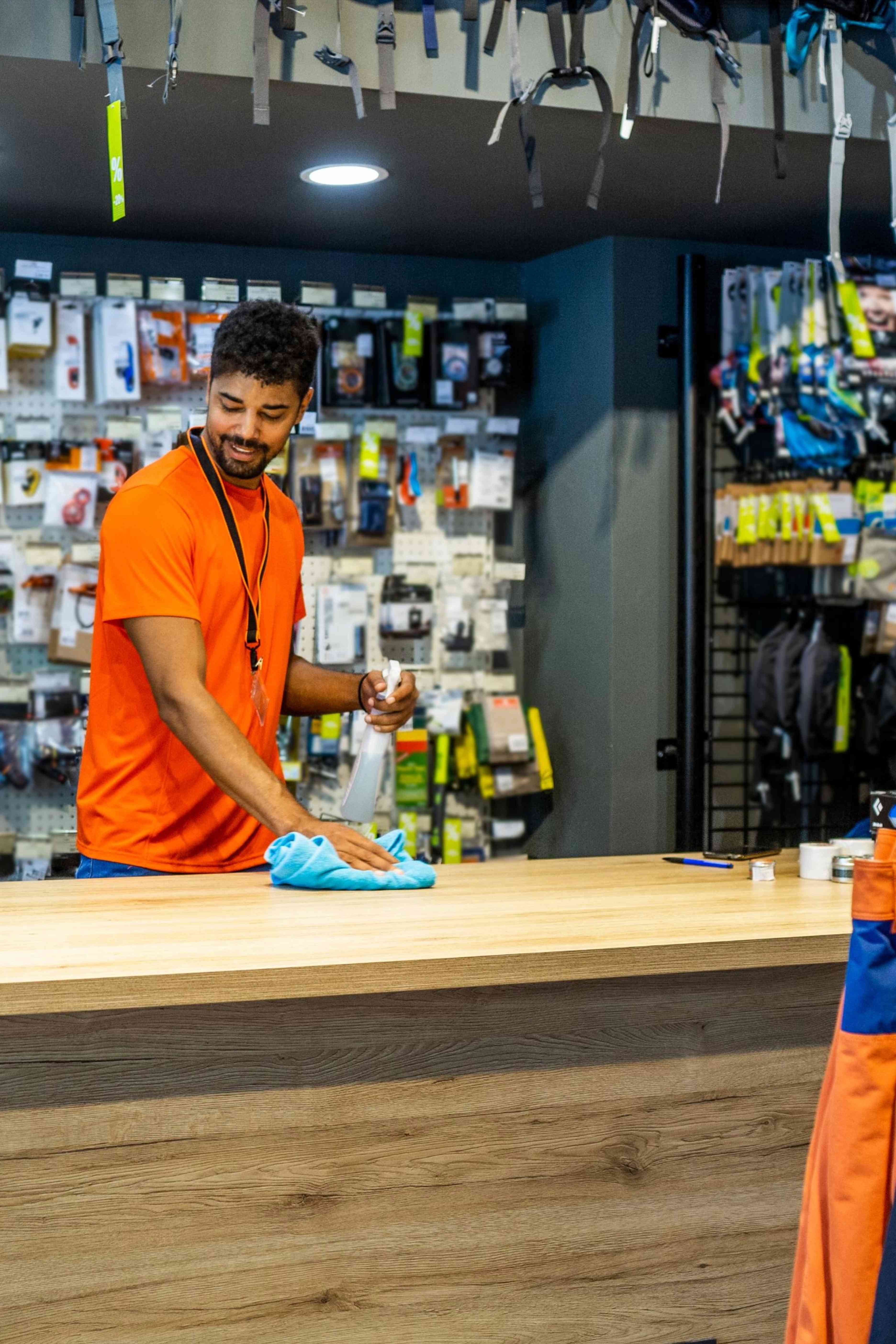 A man in an orange shirt cleaning a wooden counter with a blue cloth and spray bottle in a store.
