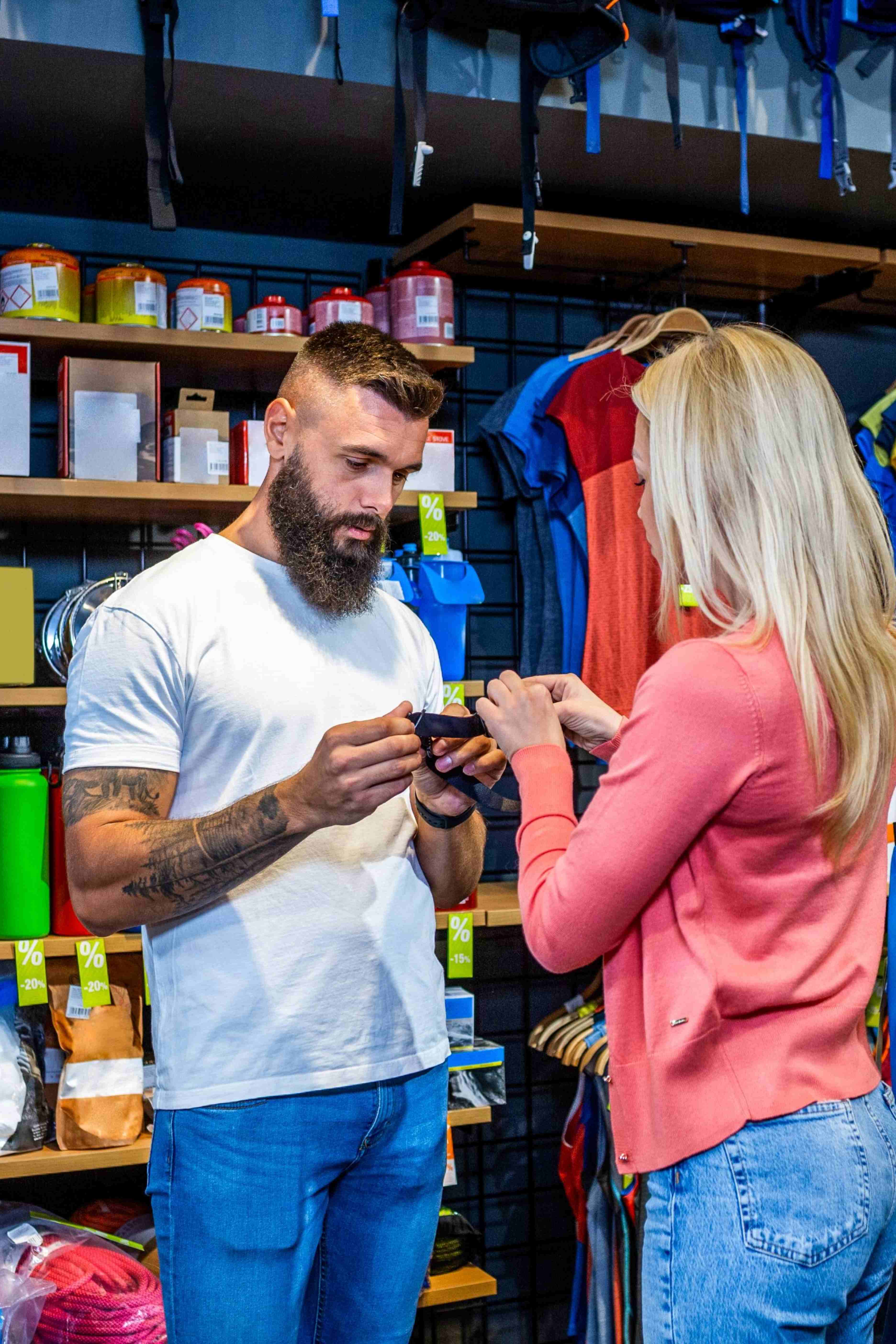 Man with beard and tattoos assists woman with blonde hair in handling a black strap in a sports or outdoor gear store.