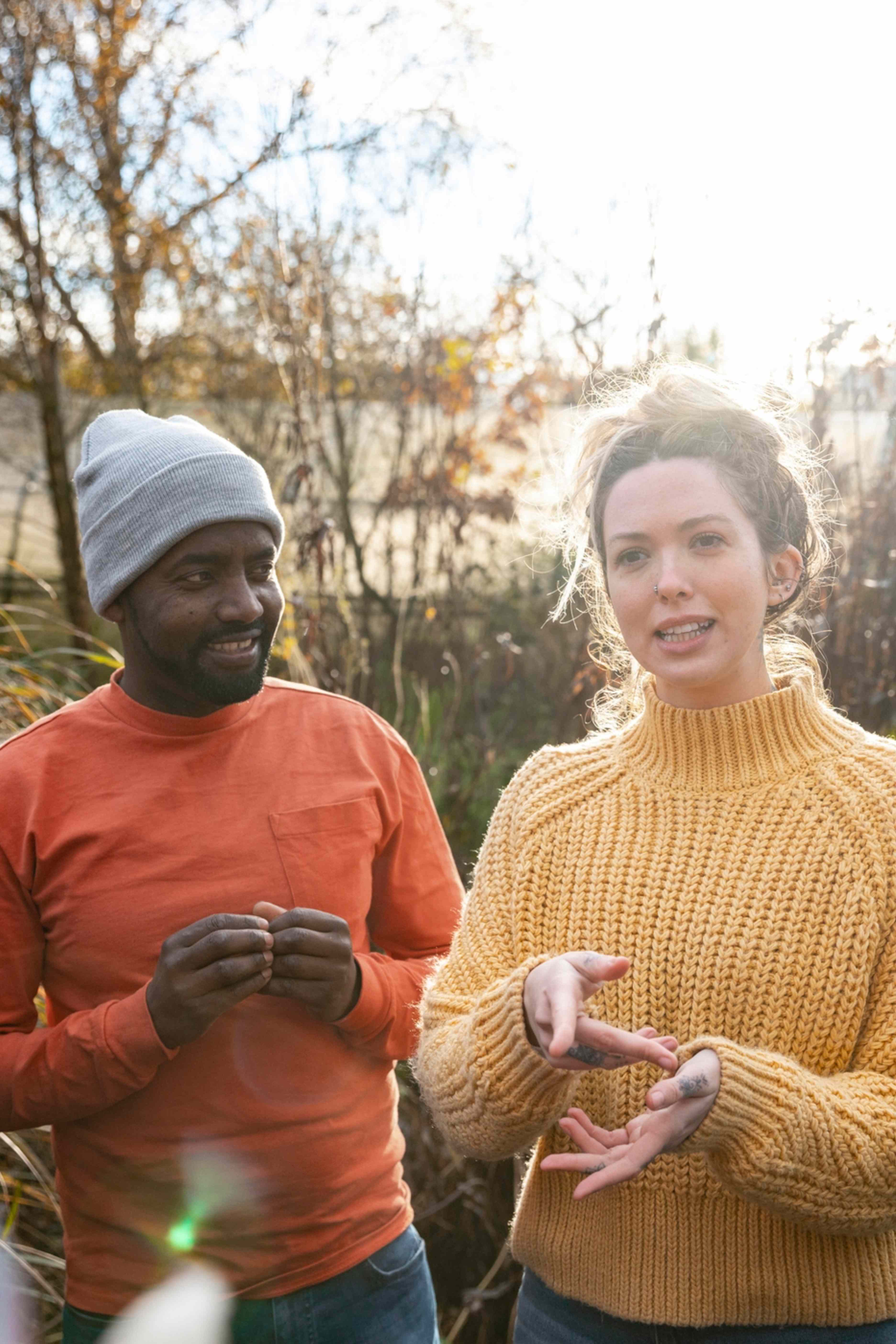 Man in orange shirt and gray beanie listens as woman in yellow sweater gestures while talking outdoors.