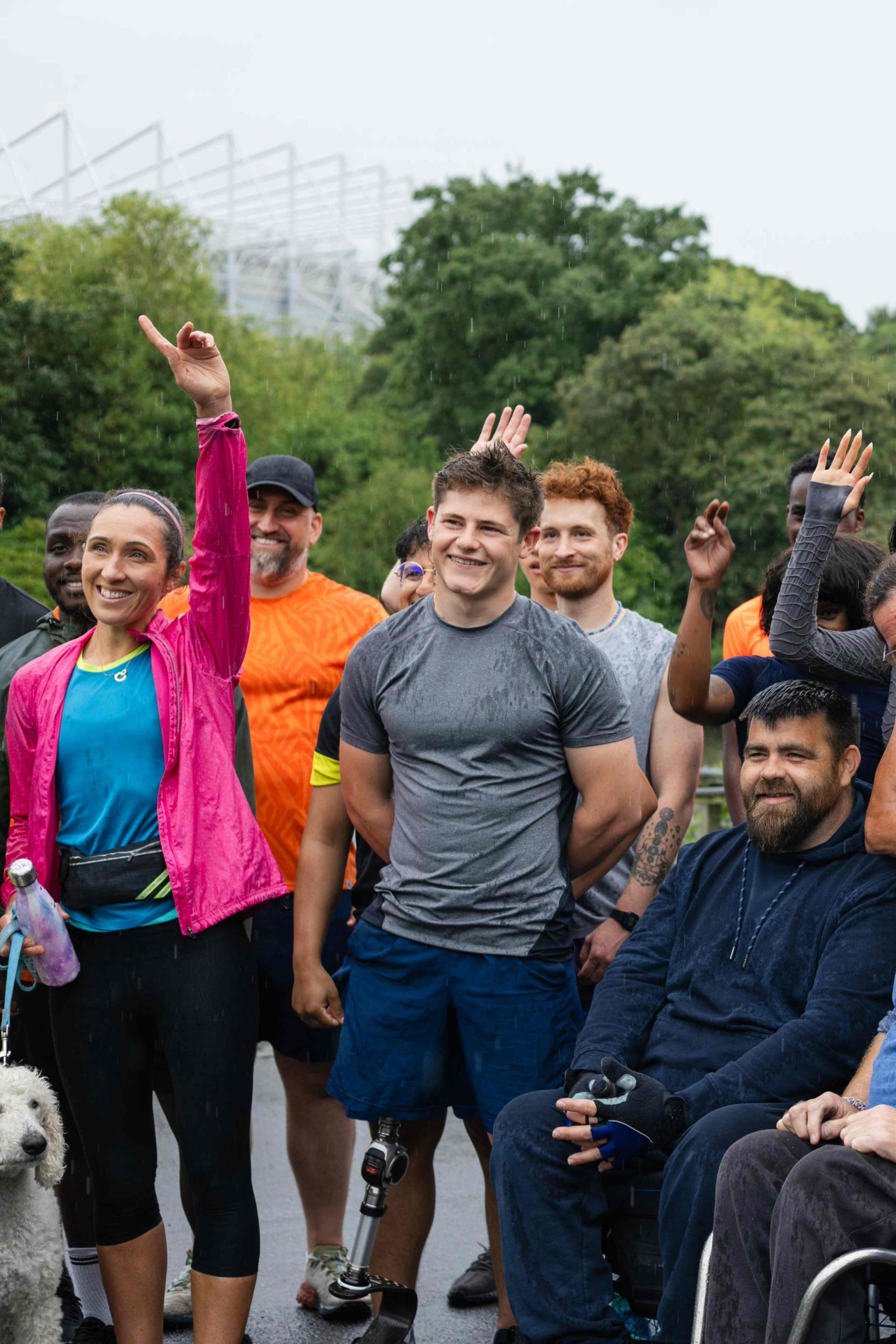 Smiling diverse group of people outdoors in casual athletic wear, including a man with a prosthetic leg and a seated man in a wheelchair, some raising their hands.