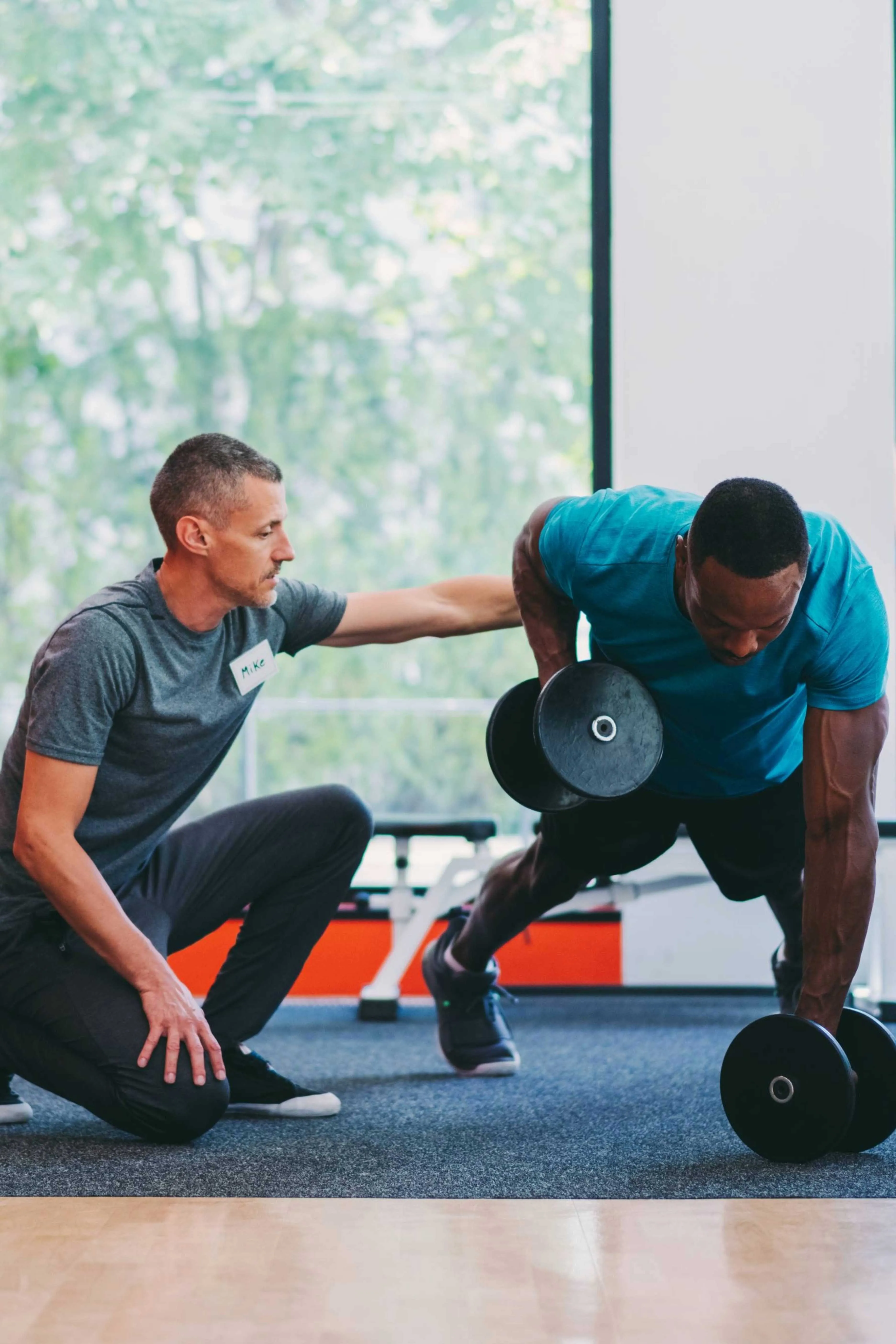 A fitness trainer wearing a name tag exercises with a man lifting dumbbells in a gym.