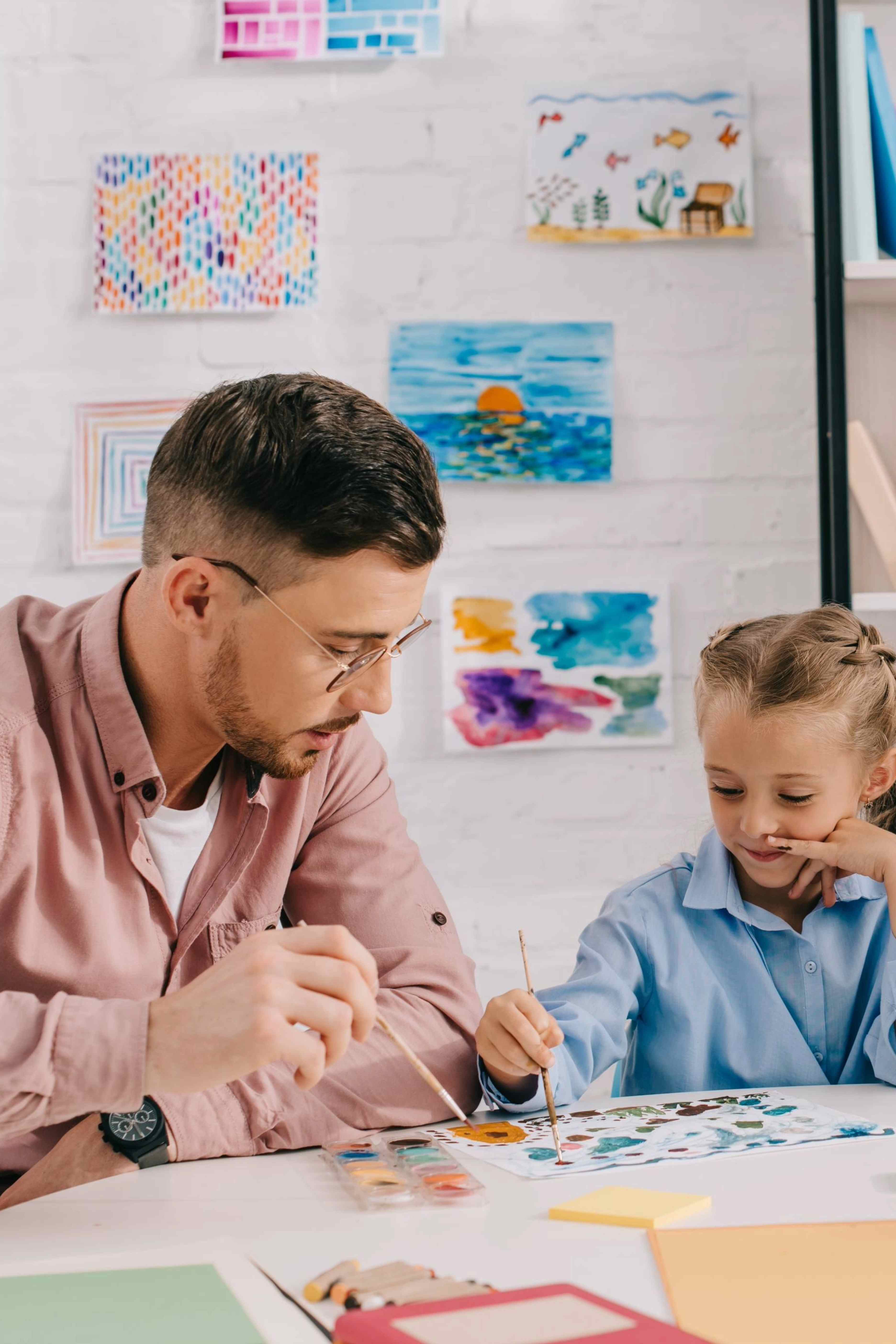 Man and young girl painting together at a table with colorful artwork on the wall behind them.