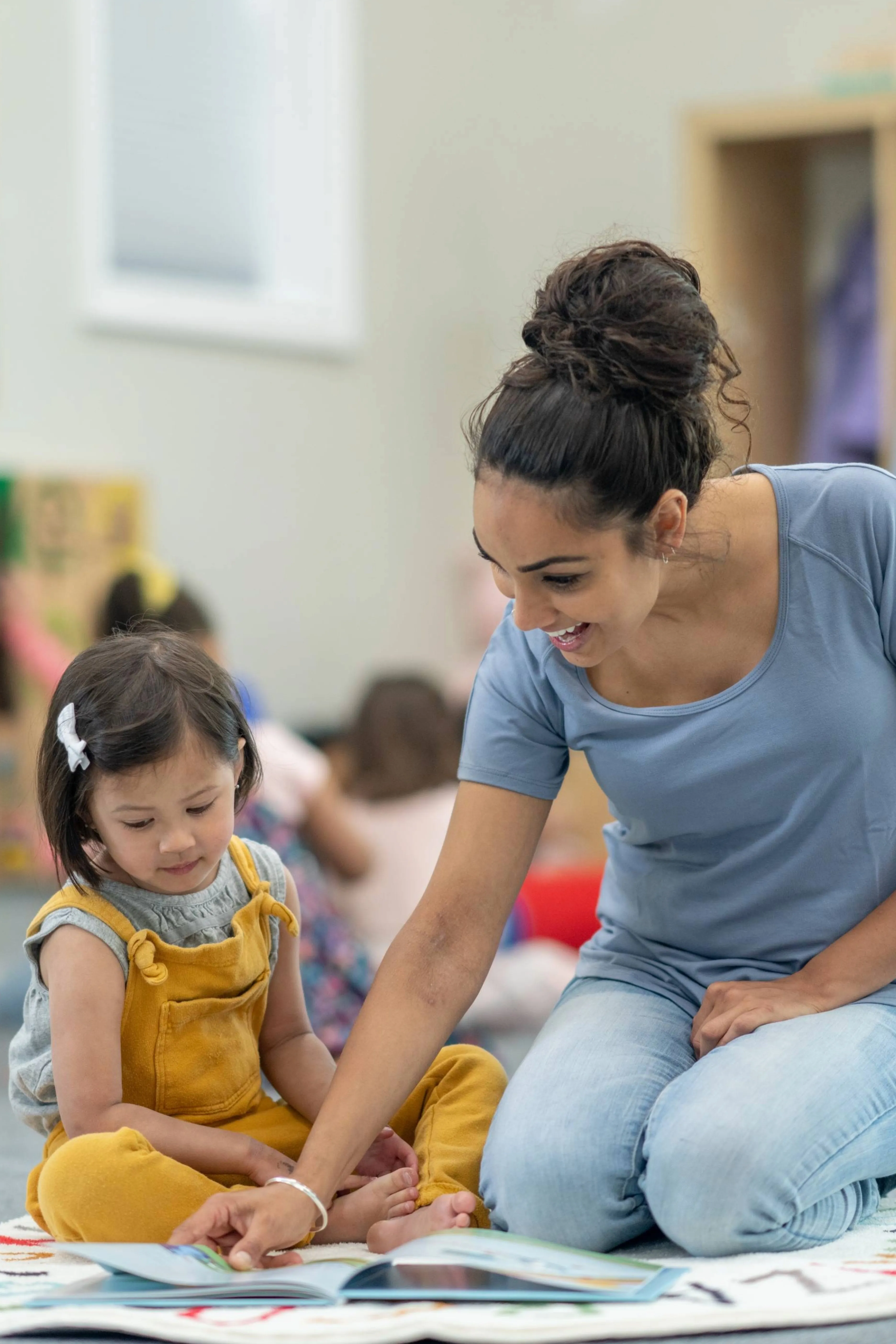 A woman and a young girl sitting on the floor reading a book together in a classroom setting.