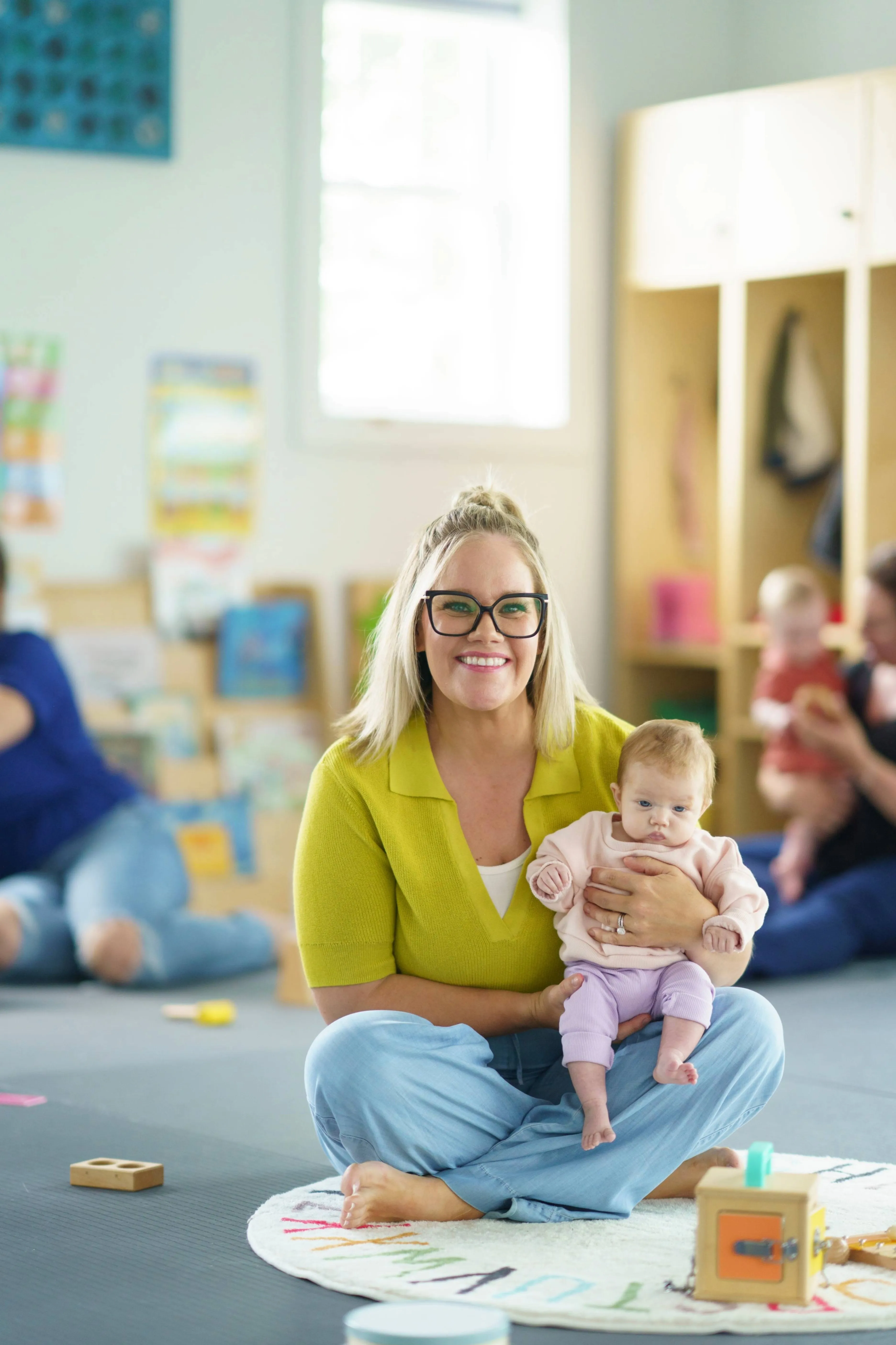 Smiling woman with glasses sitting cross-legged on a play mat holding a baby in a bright room with toys and other adults and children in the background.