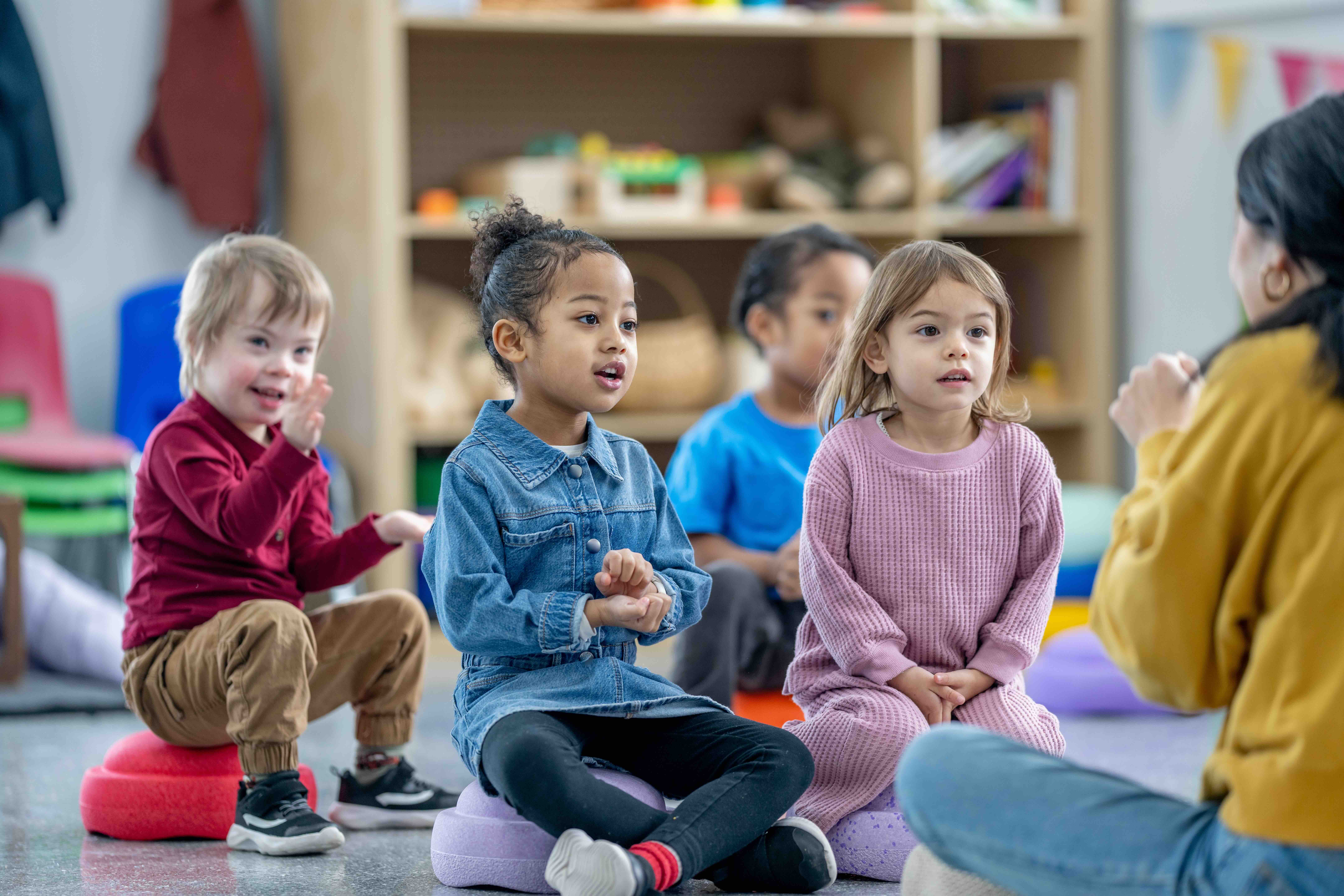 Four young children sitting on colorful cushions in a classroom, engaging with a teacher.