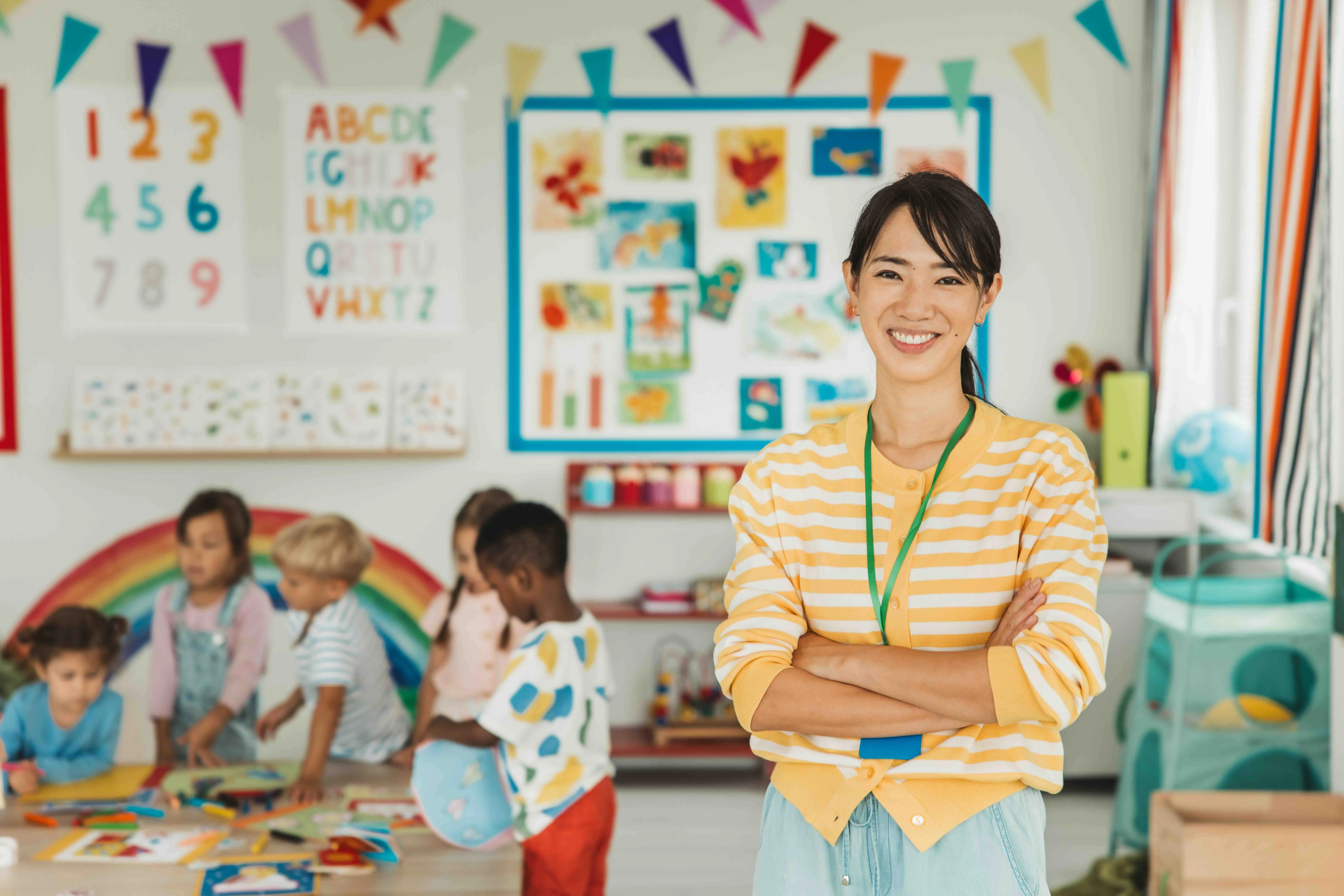 Smiling female teacher in a yellow striped sweater standing with arms crossed in a colorful classroom with children playing at a table.