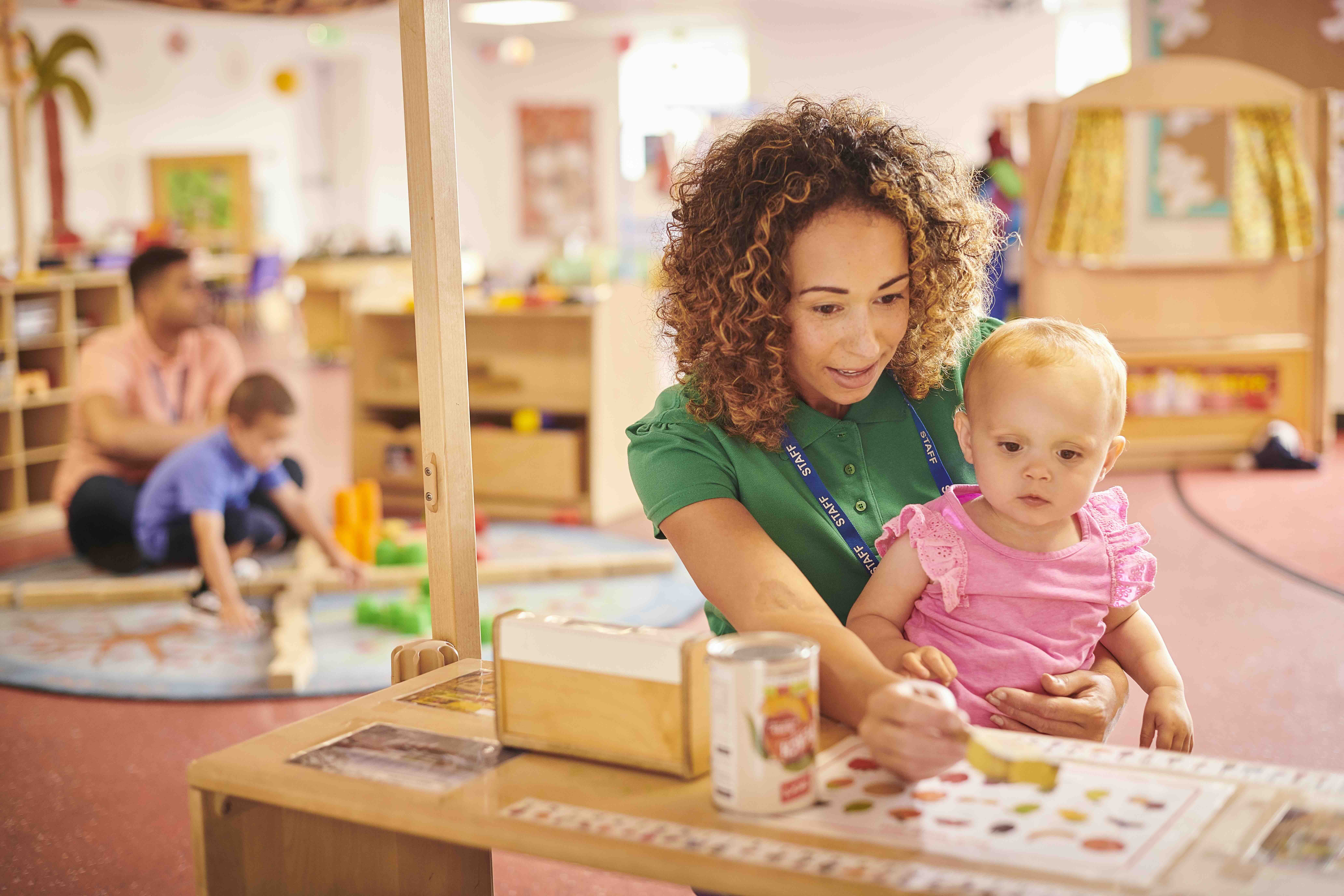 Childcare worker in a green shirt showing a young toddler in pink a picture chart in a colorful classroom.