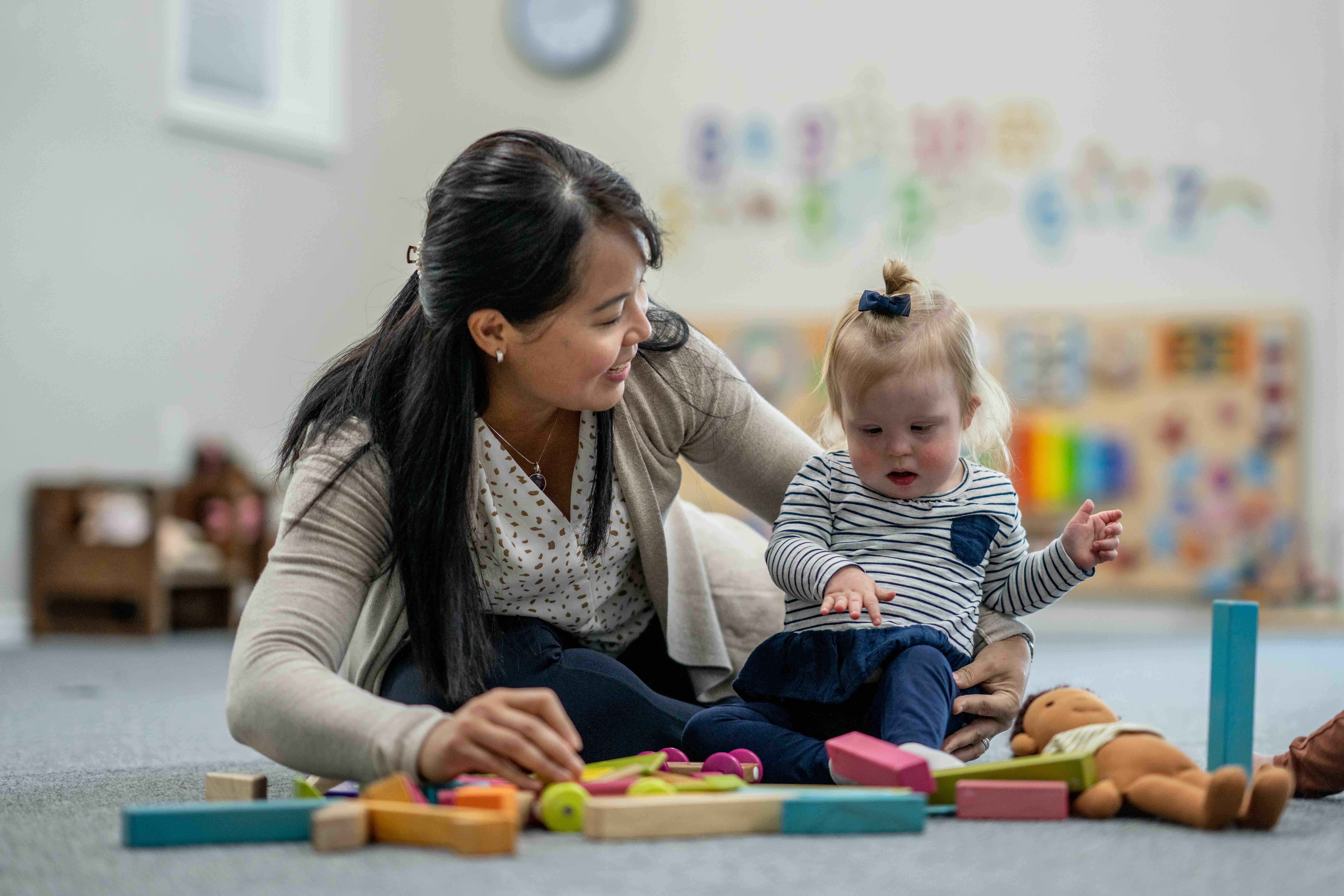 Woman playing on the floor with a toddler surrounded by colorful wooden blocks and a doll in a classroom setting.