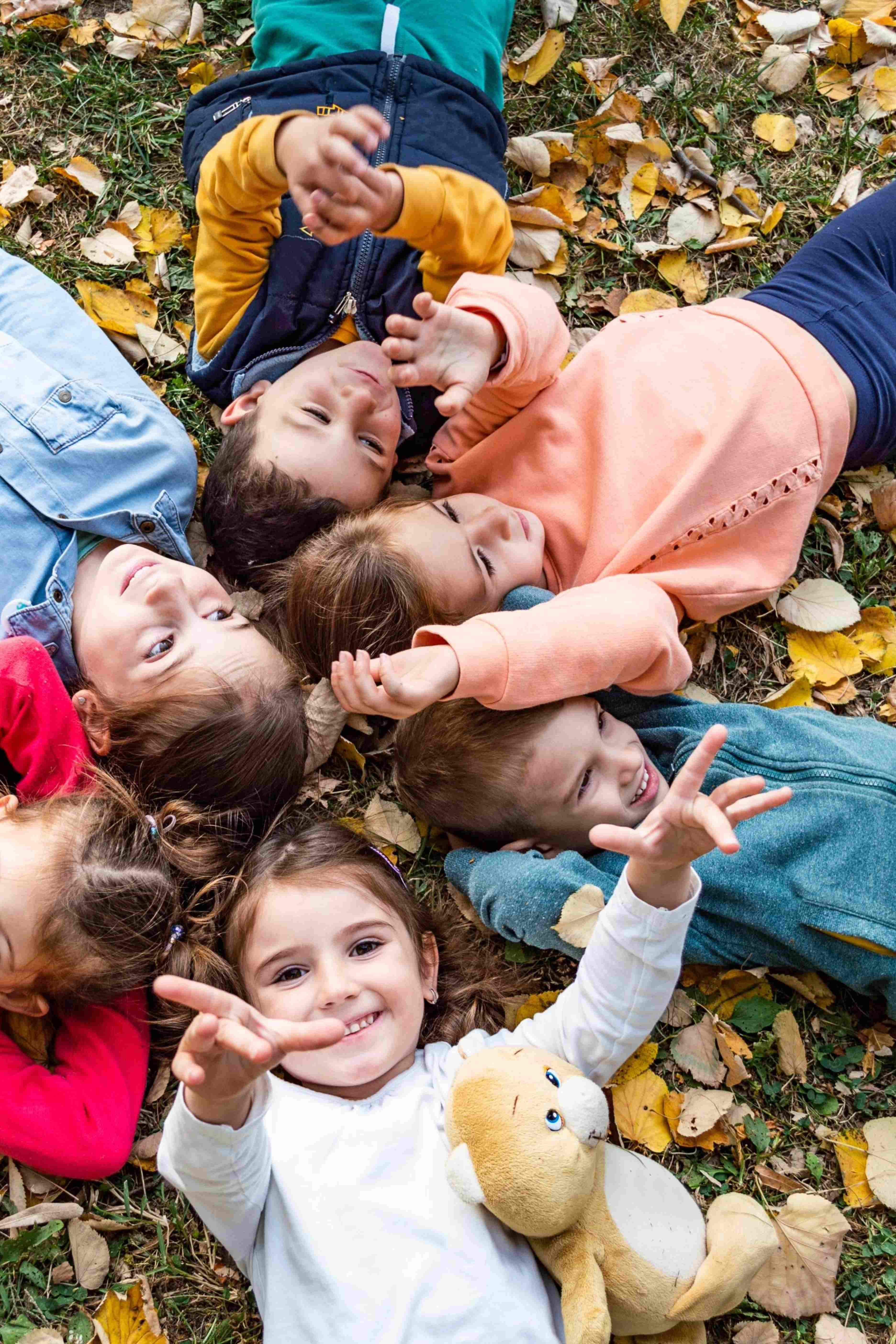 Six children lying on autumn leaves in a circle, smiling and reaching towards the camera, one holding a teddy bear.