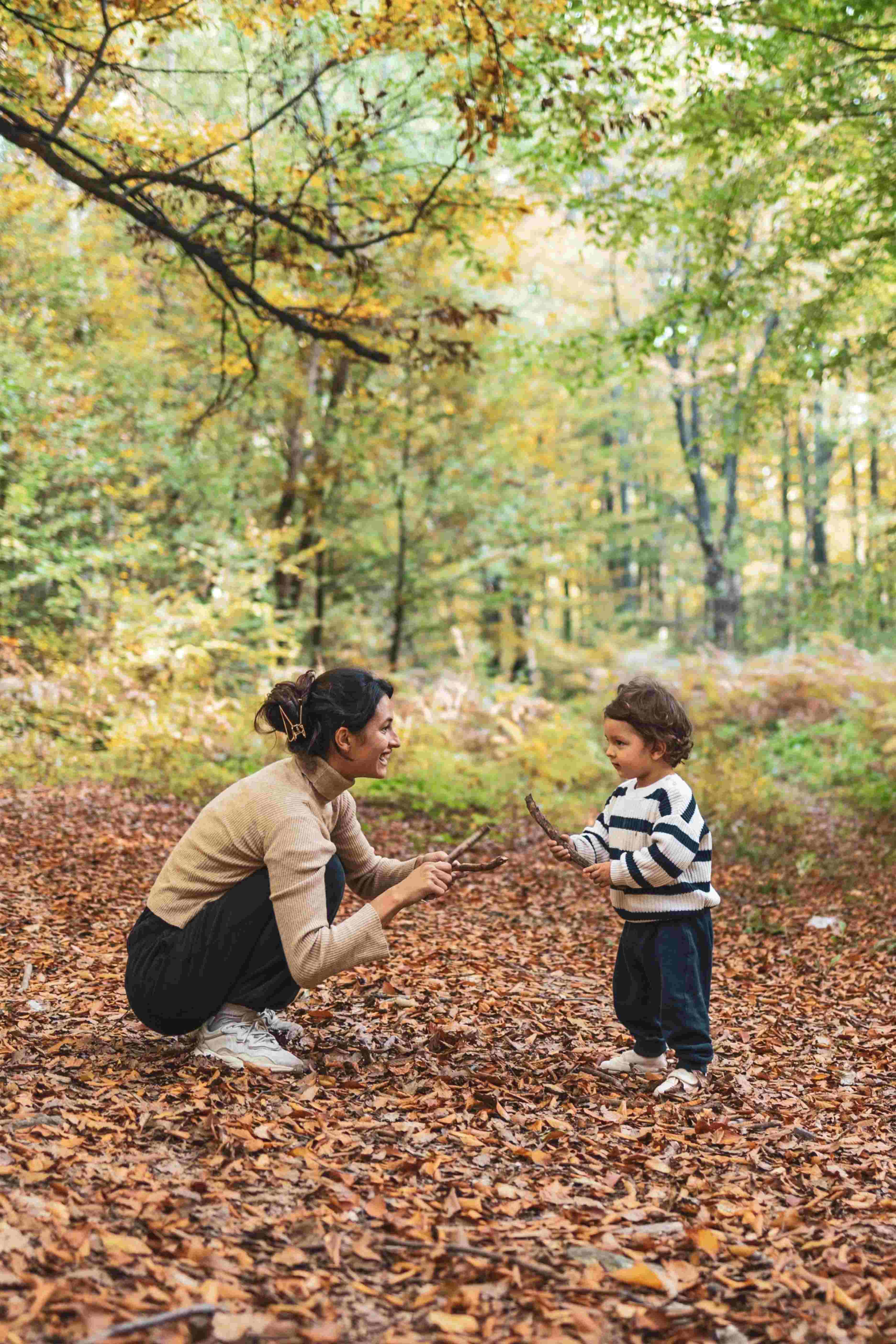 A woman squatting and smiling while holding sticks, playing with a toddler standing on a leaf-covered forest ground.