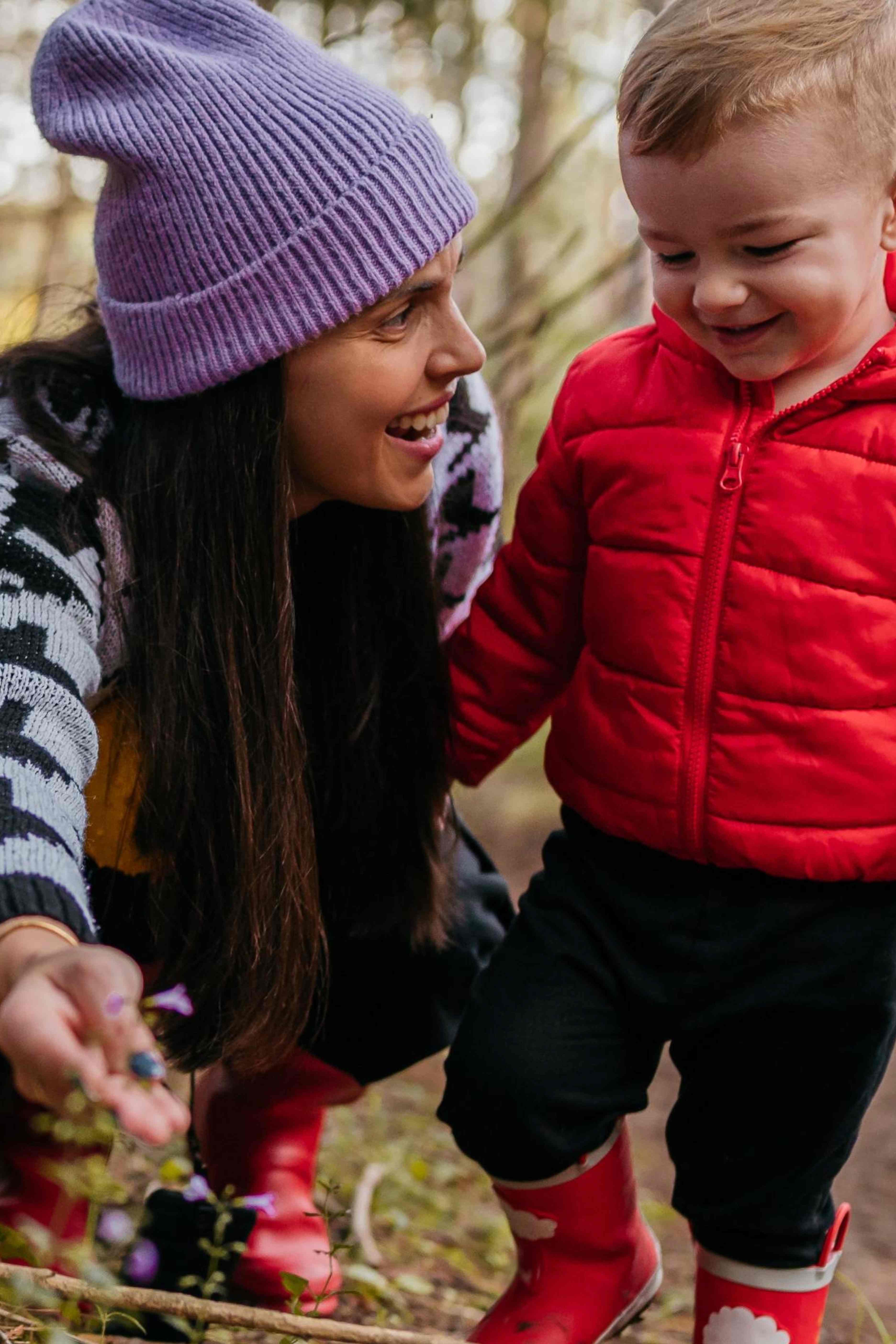 Woman in a purple beanie and patterned sweater kneels smiling and showing a small flower to a toddler in a red jacket and red rain boots outdoors.