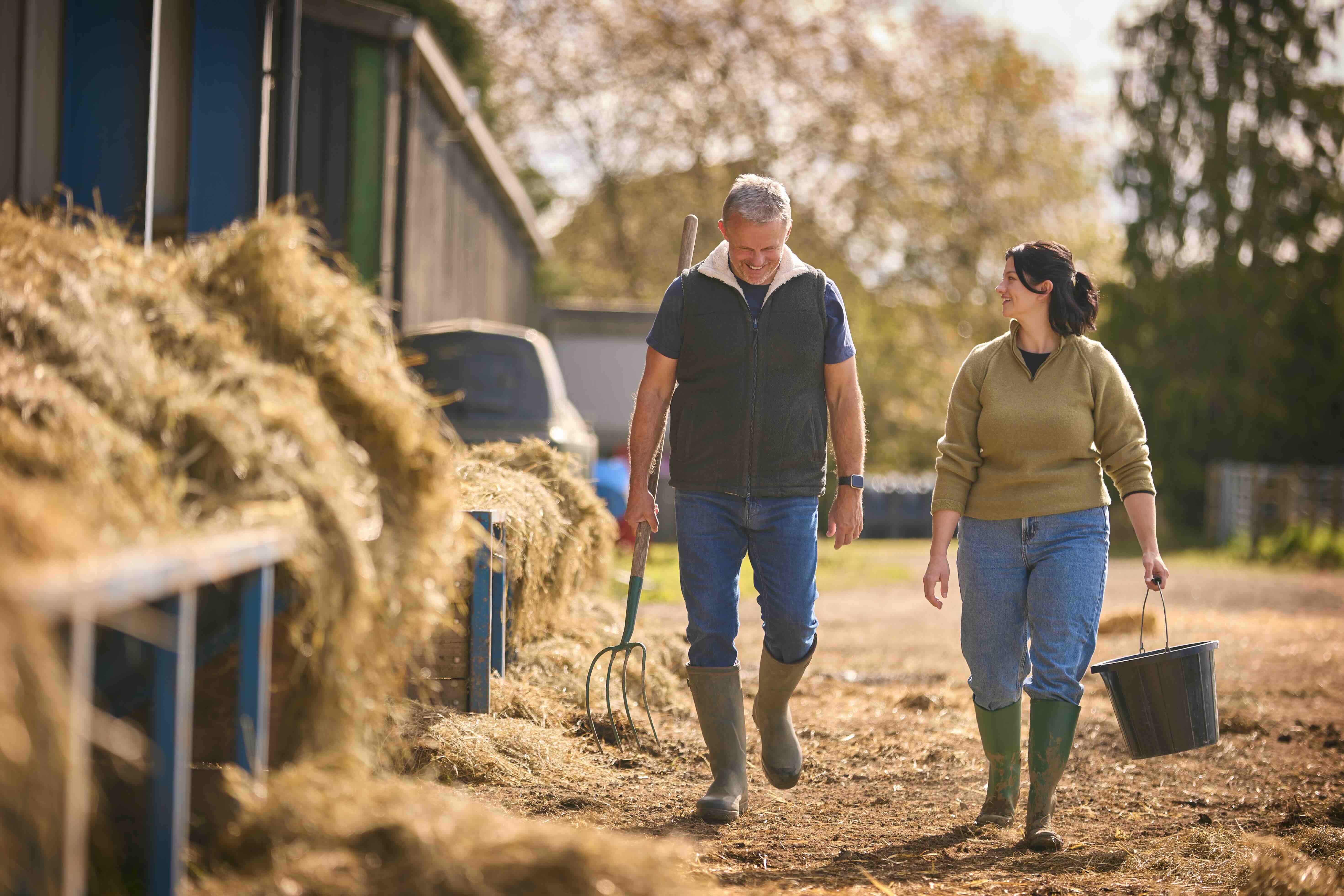 Two farmers walking outdoors carrying a pitchfork and a black bucket, surrounded by hay and farm buildings.