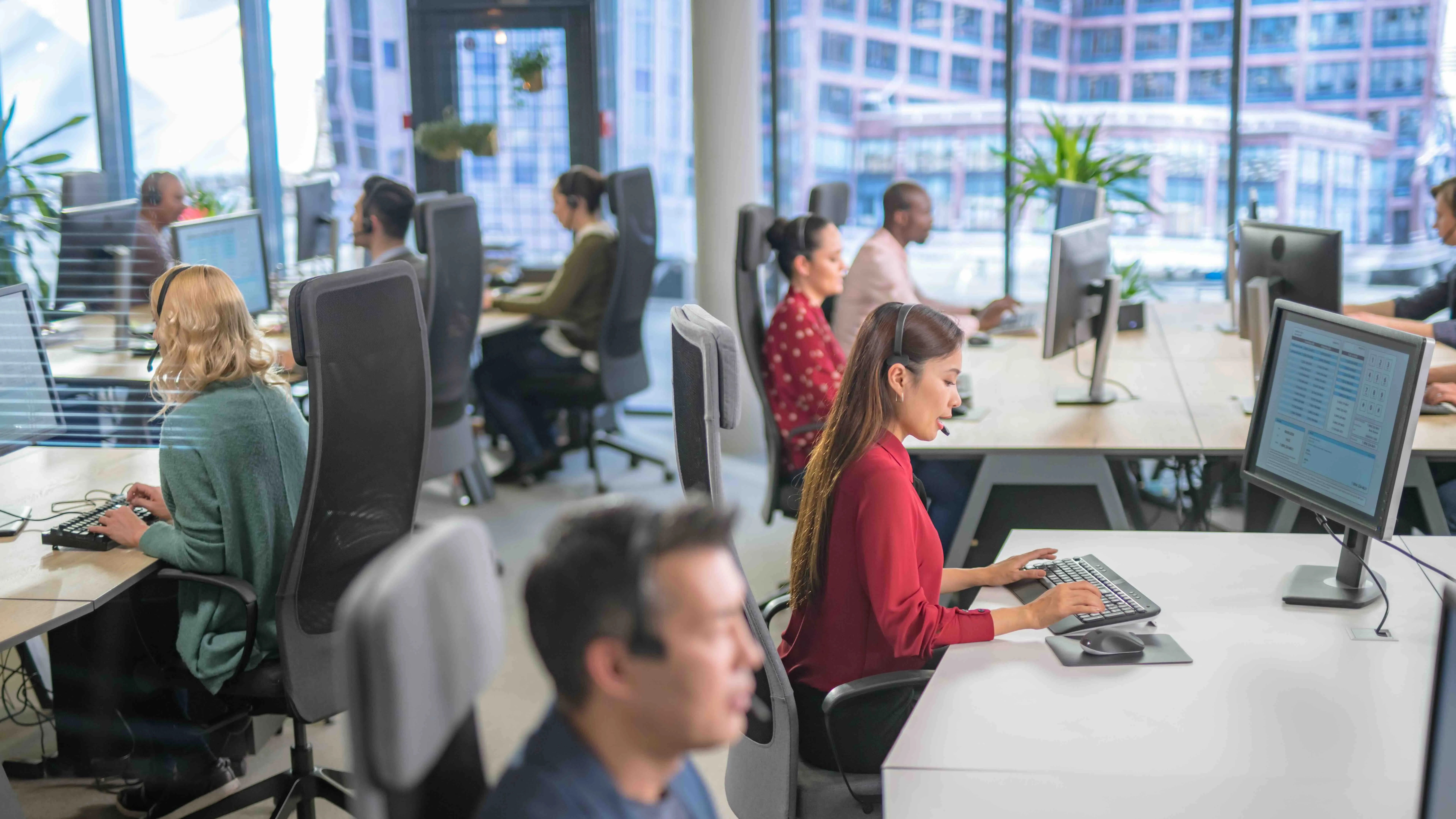 Customer service representatives wearing headsets working at their computers in a modern office with large windows.