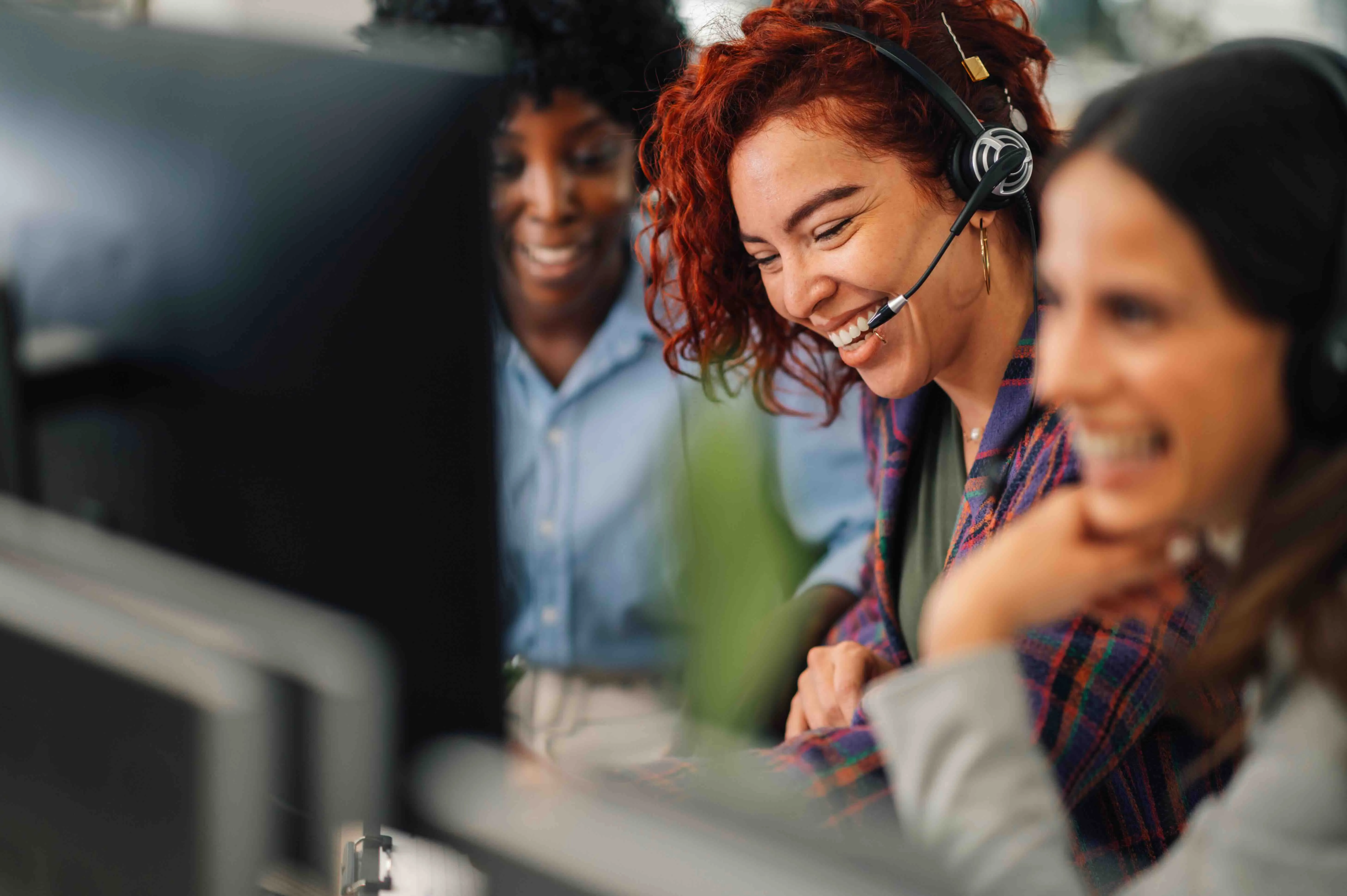 Three smiling women wearing headsets collaborating in an office environment.