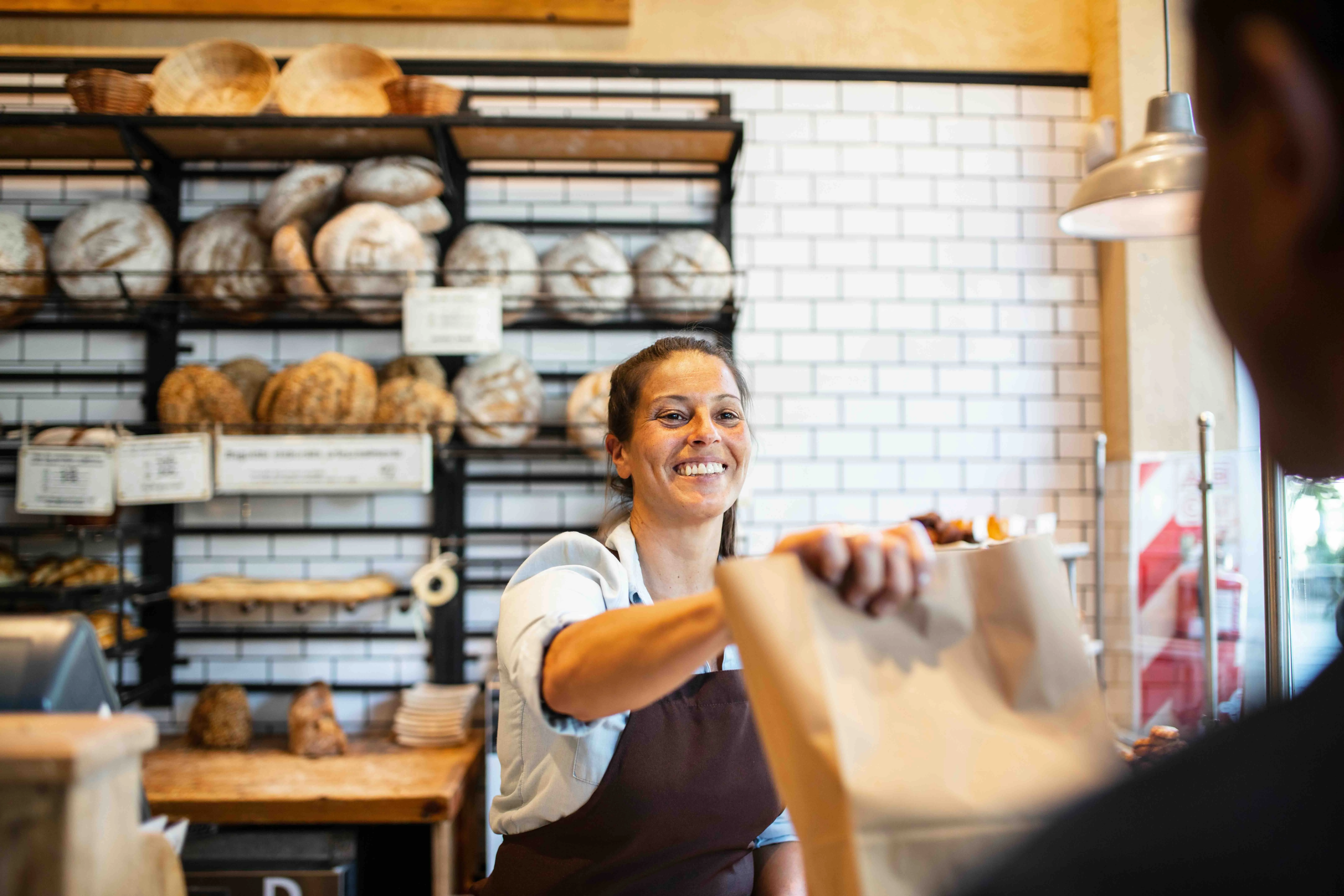 Smiling bakery worker handing a paper bag to a customer with shelves of bread in the background.