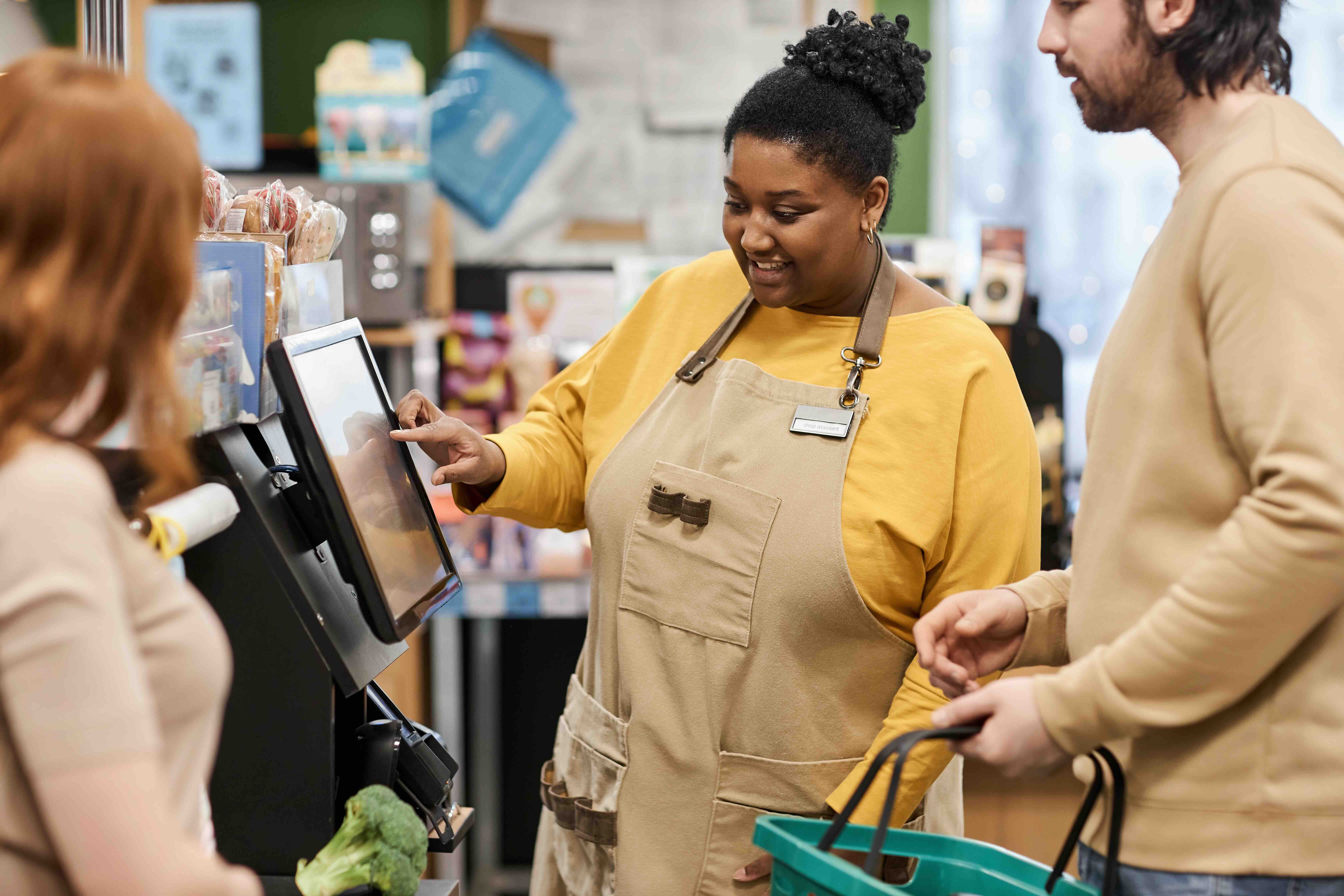 Grocery store employee wearing an apron assisting two customers at a touchscreen self-checkout kiosk.