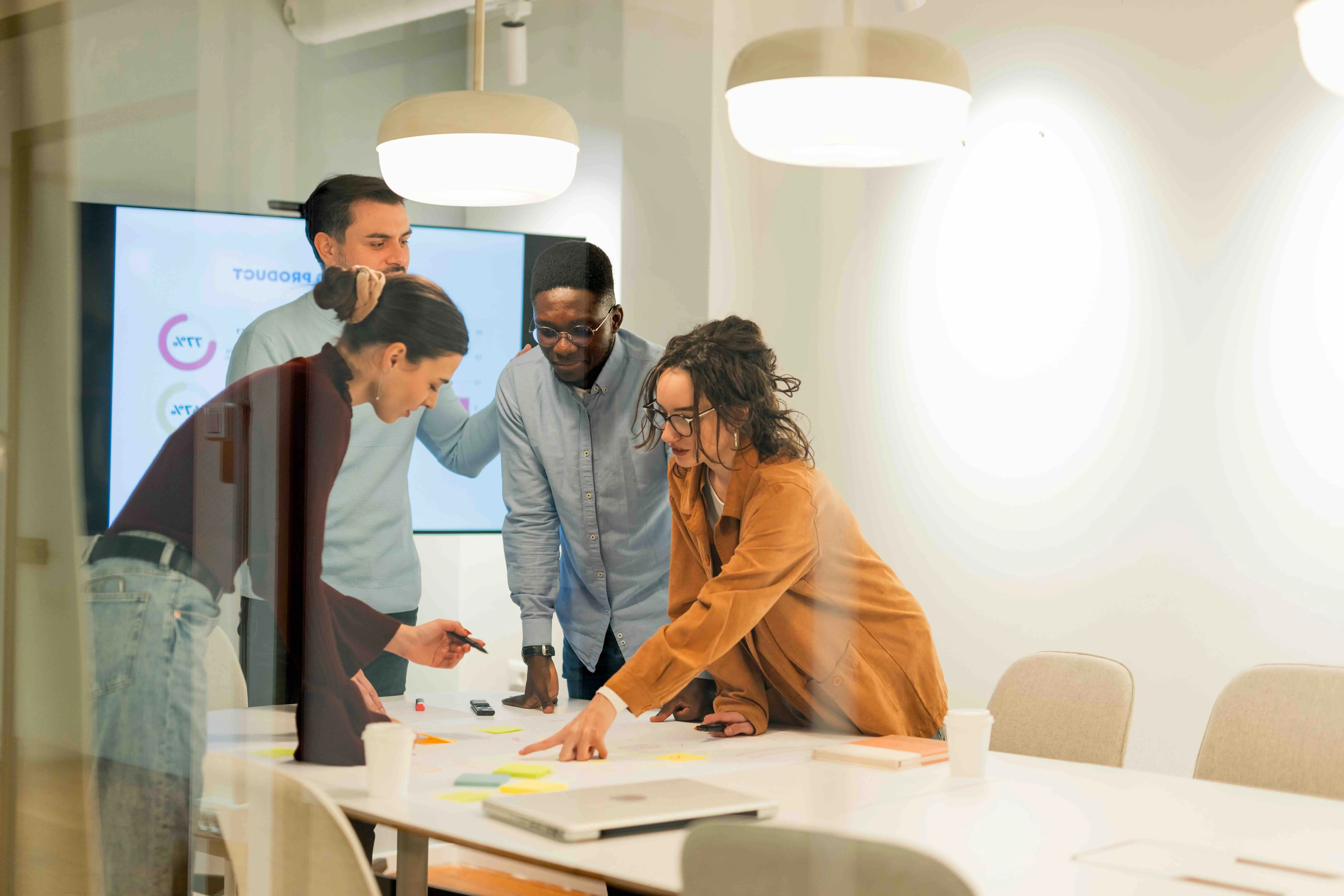 Four diverse colleagues collaborating around a table with sticky notes and laptop in a bright conference room.