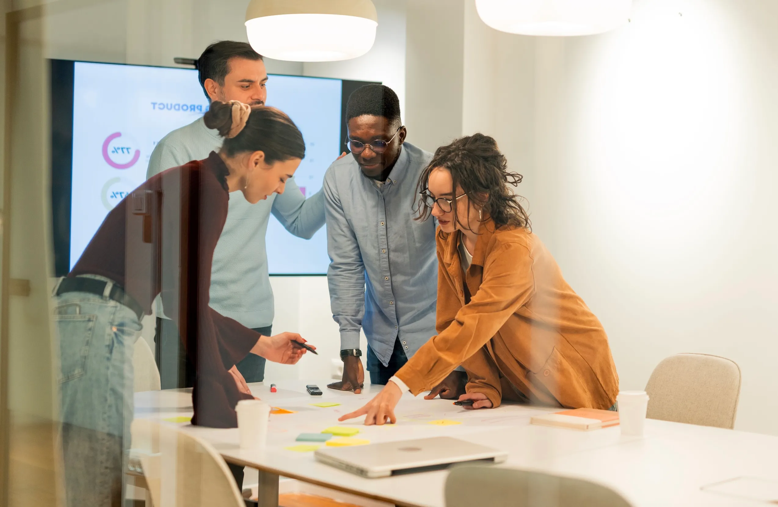 Four young professionals collaborating over a table with sticky notes and documents in a modern office meeting room.