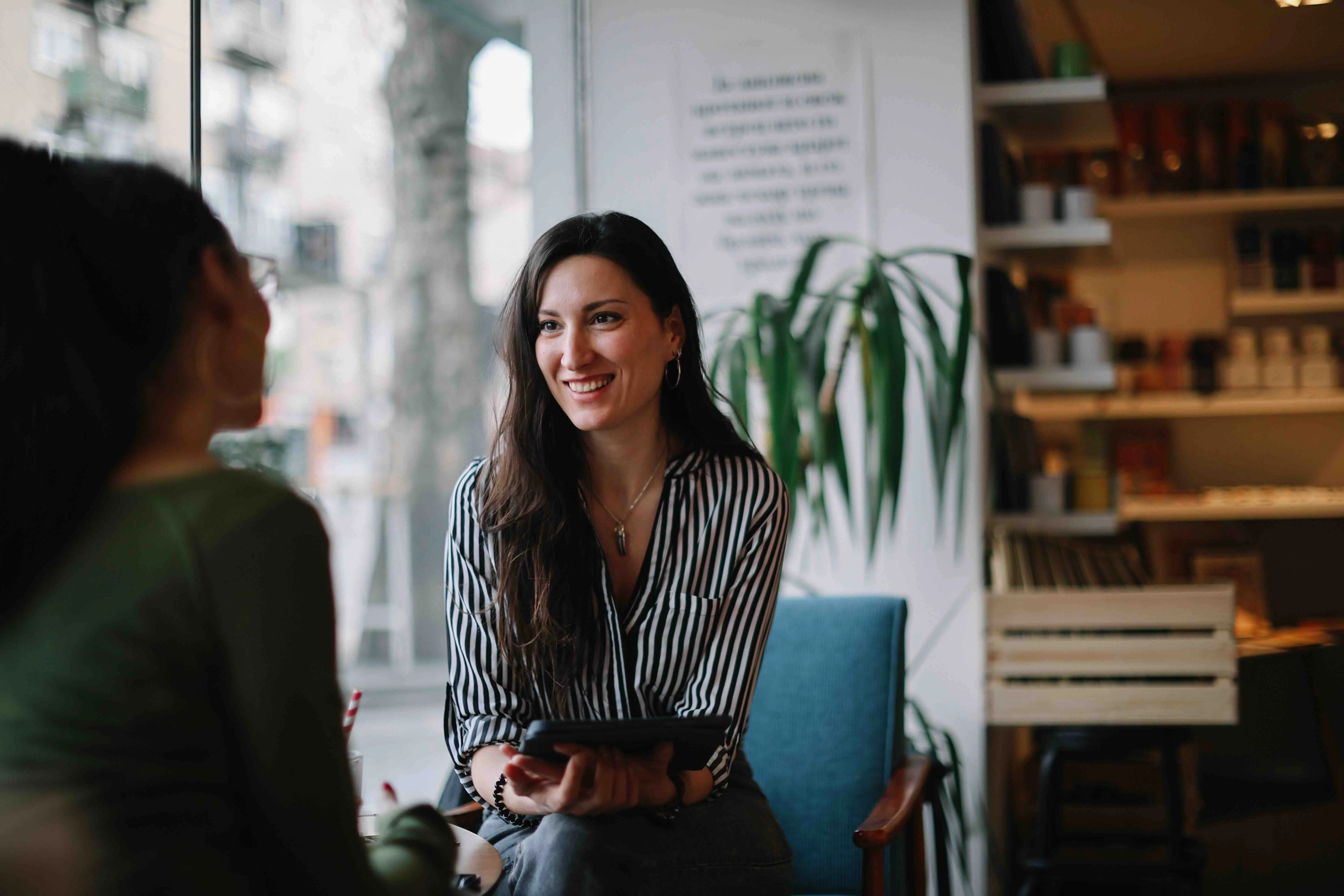 Two women having a friendly conversation in a cozy cafe, one holding a tablet and smiling.