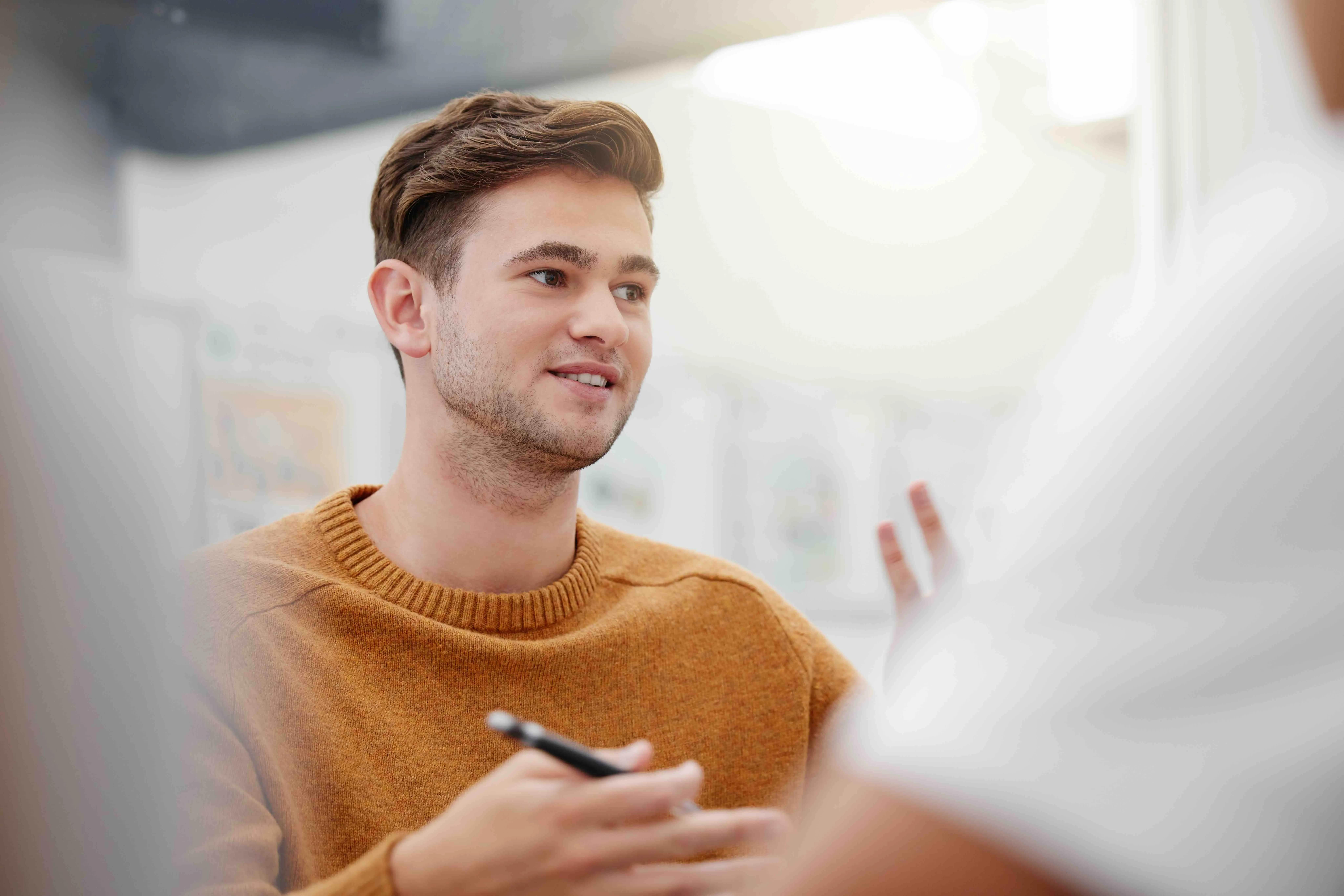Young man in a mustard sweater engaging in a conversation while holding a pen.