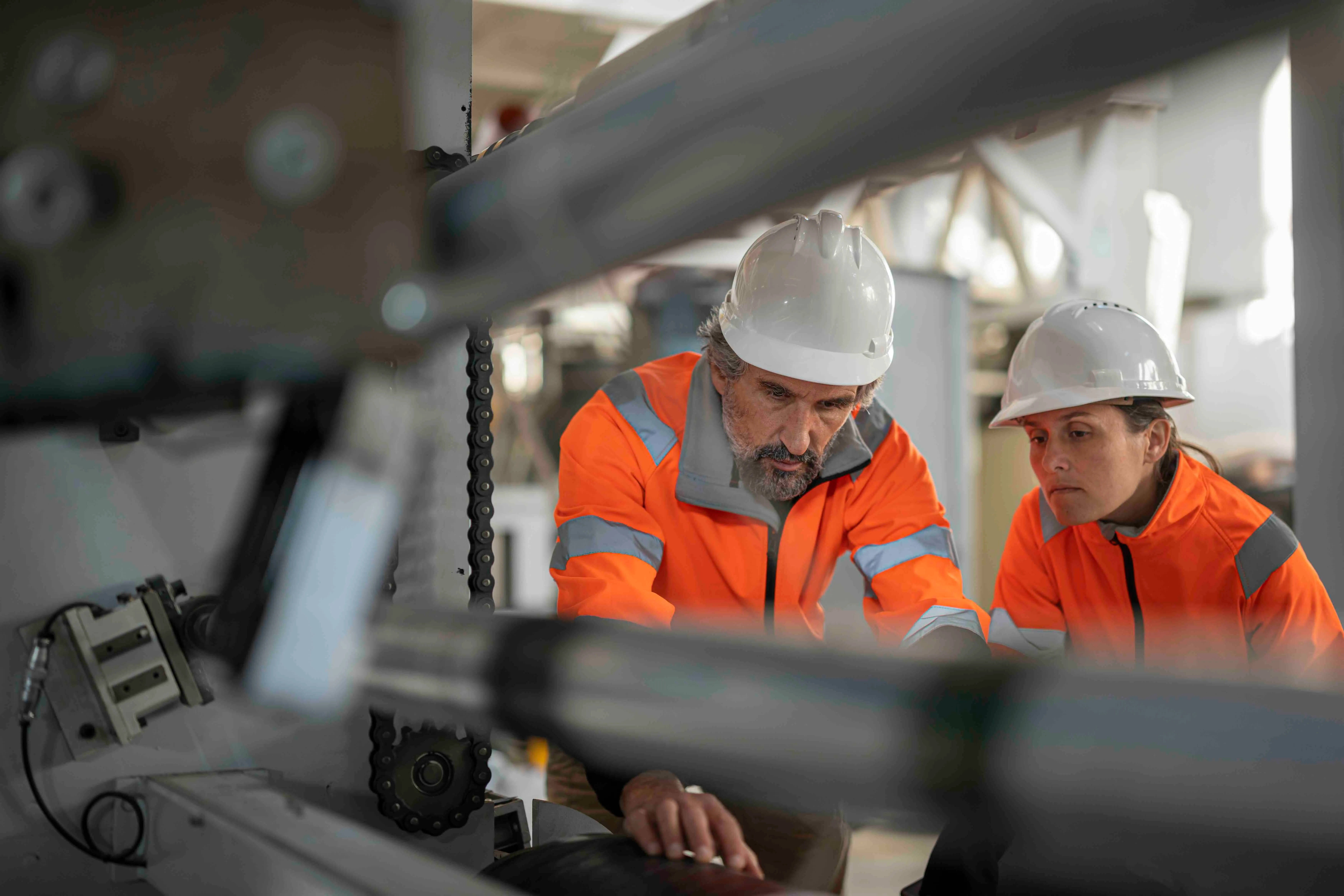 Two workers wearing white hard hats and orange safety jackets inspecting machinery in an industrial setting.