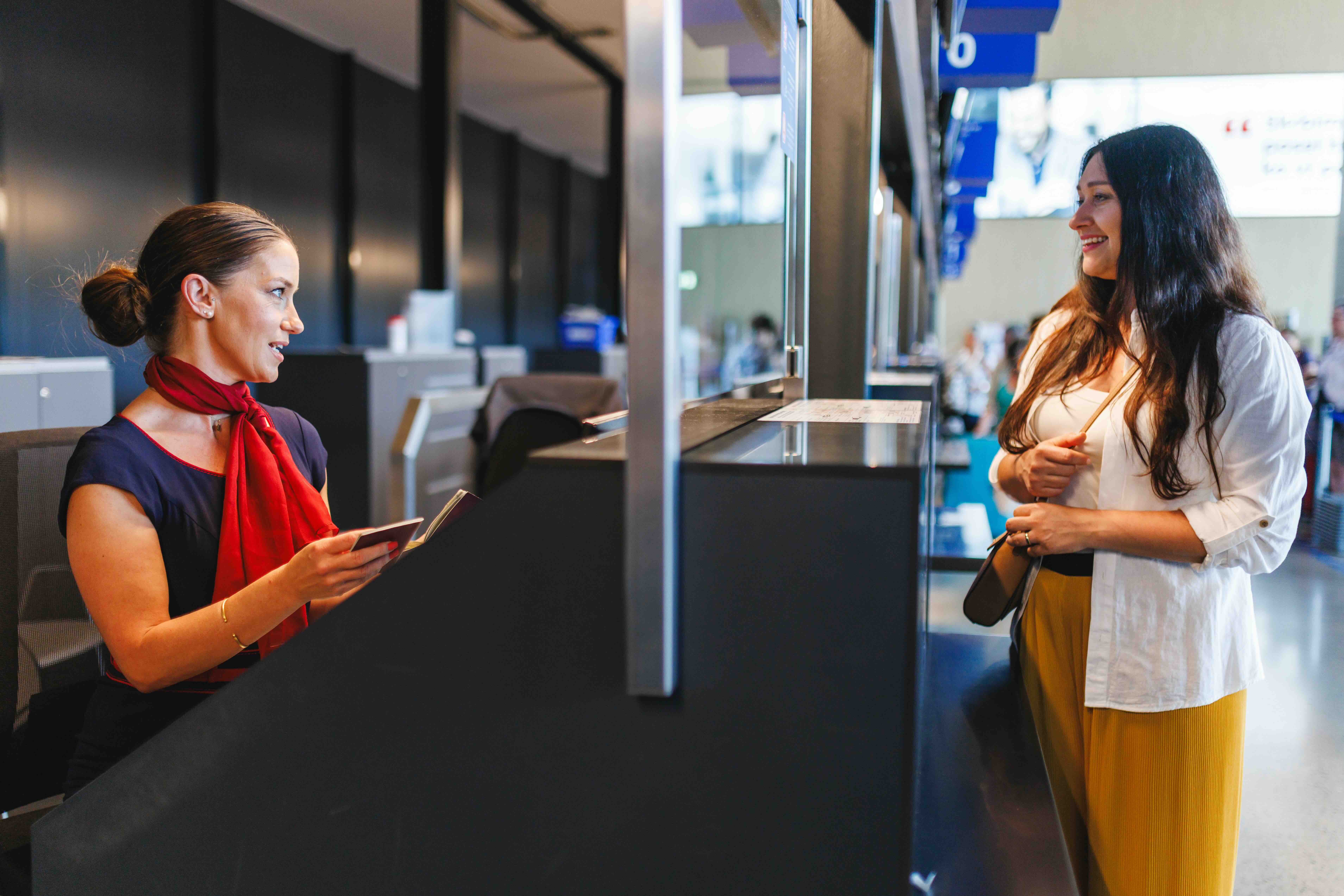 Airport check-in agent in red scarf assisting female traveler with documents at counter.