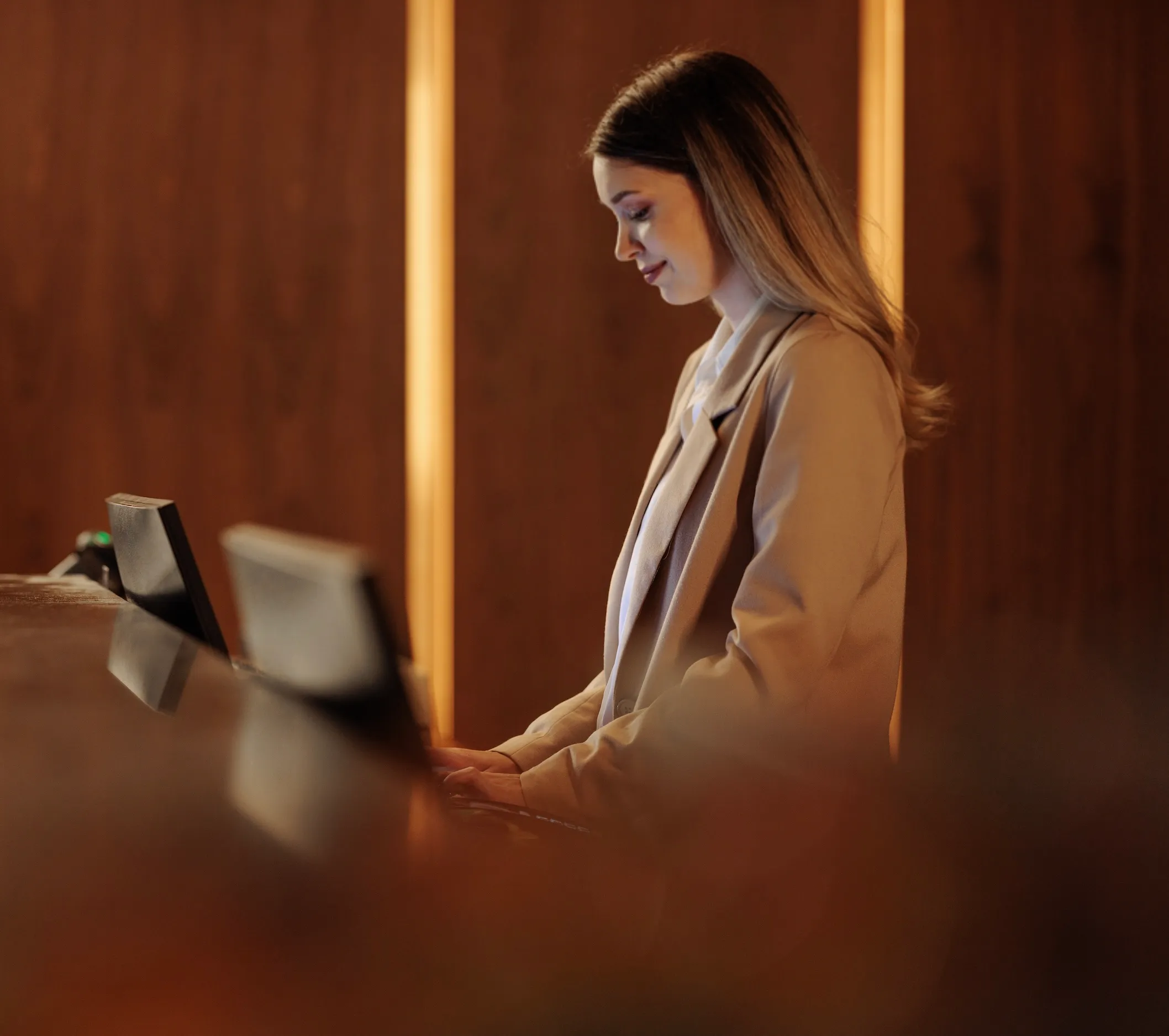 Woman standing at a hotel reception desk typing on a keyboard with warm wood-paneled walls in the background.