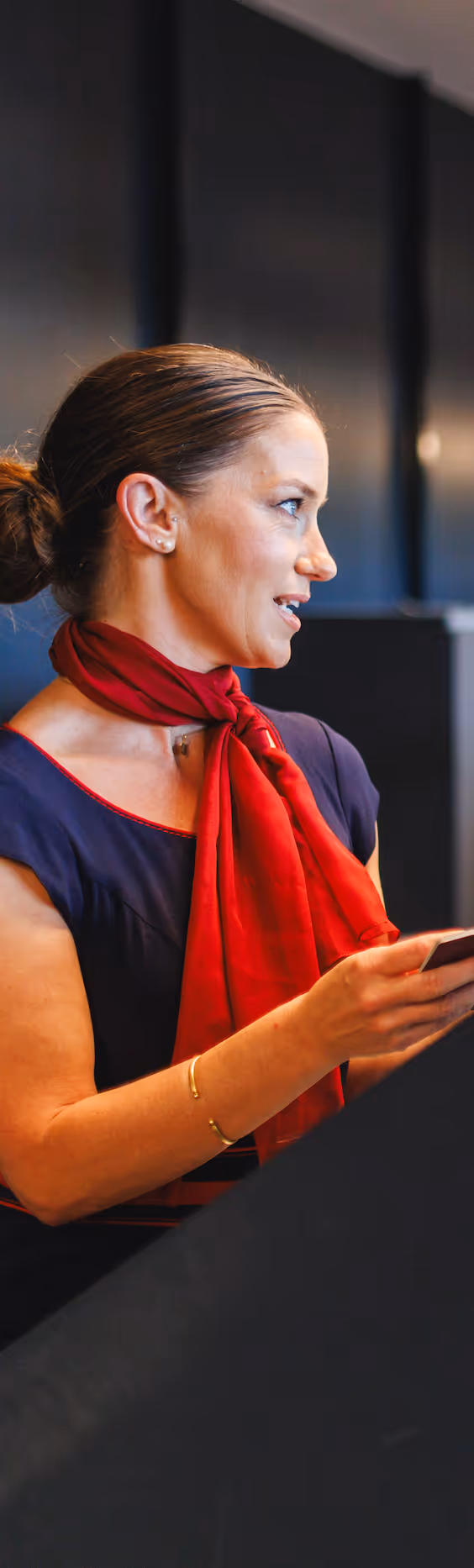 Woman with brown hair tied back wearing a red scarf and dark top, holding a smartphone and speaking.