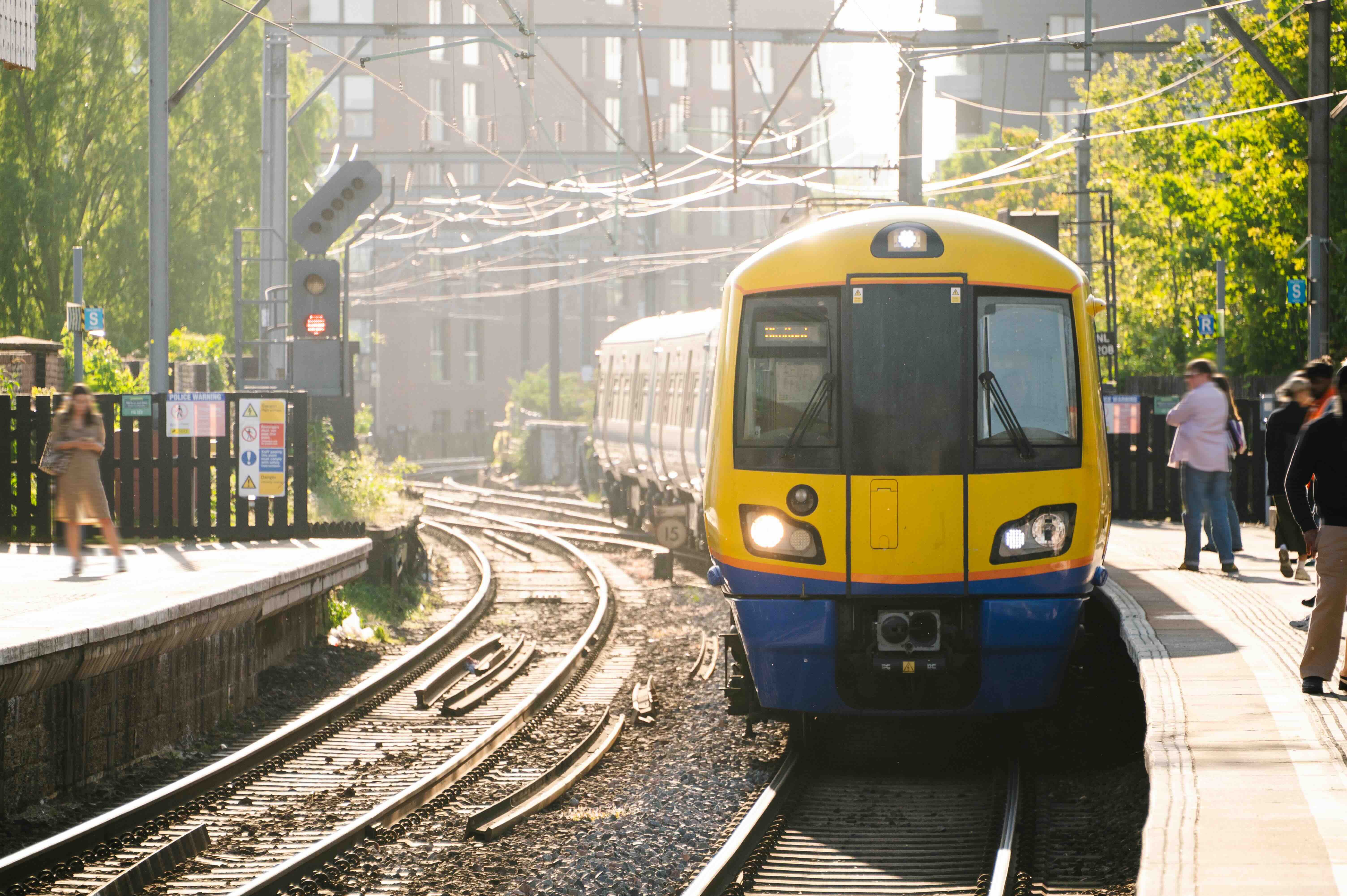 Yellow and blue commuter train approaching station platform with passengers waiting on a sunny day.