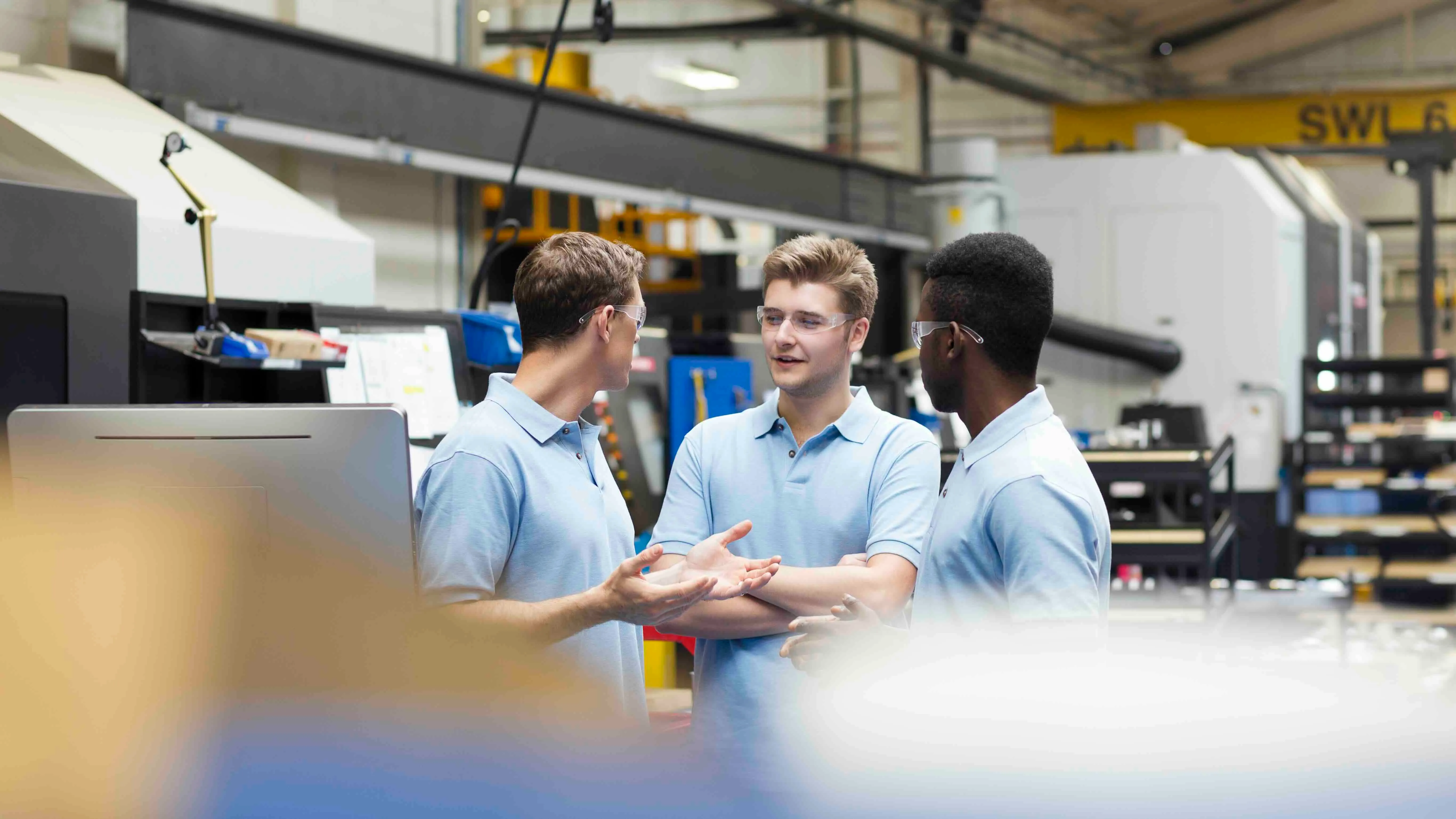 Three men wearing safety glasses and light blue polo shirts having a discussion in an industrial setting.