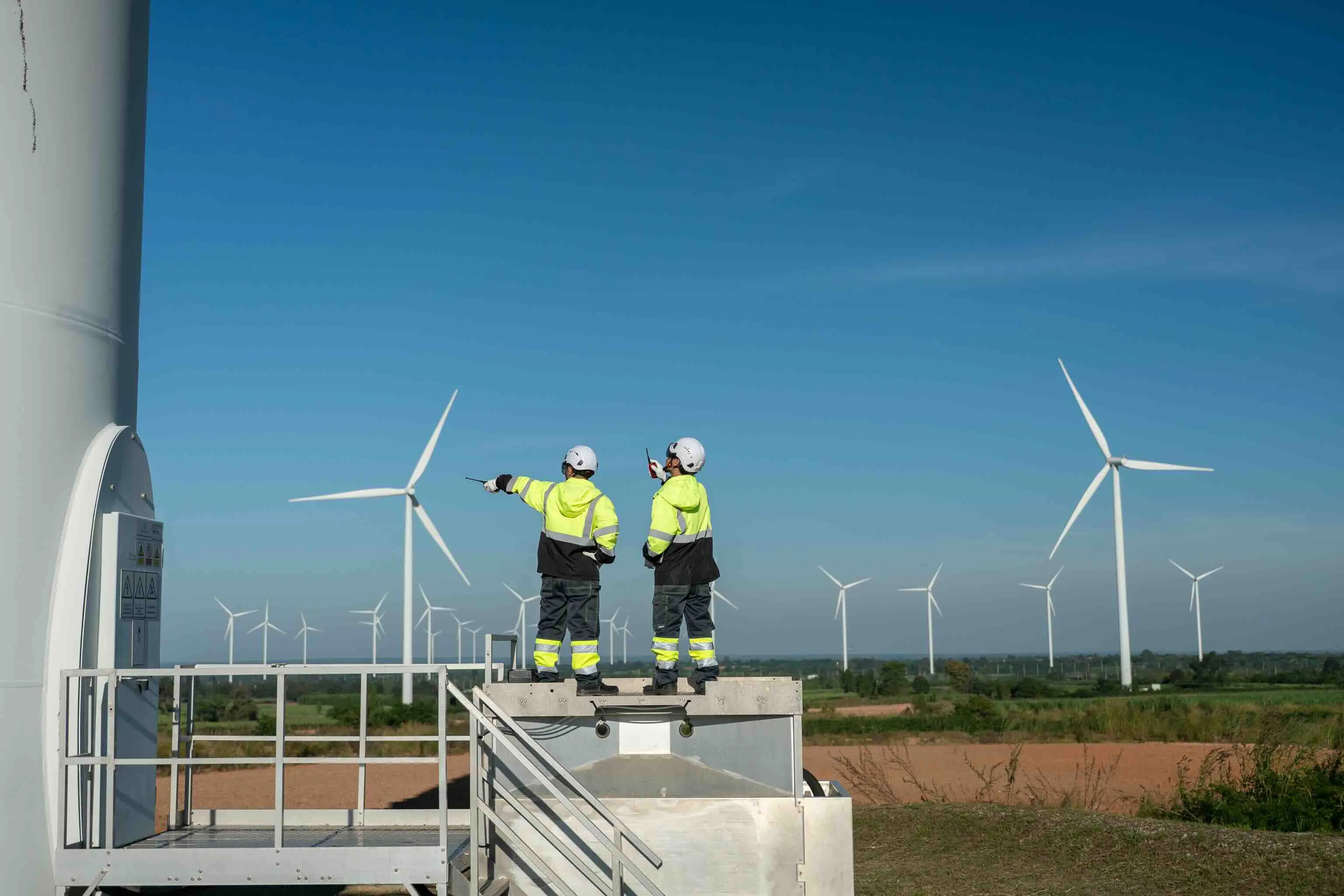Two workers in yellow and black safety jackets and helmets standing on a platform near wind turbines in a field under a clear blue sky.