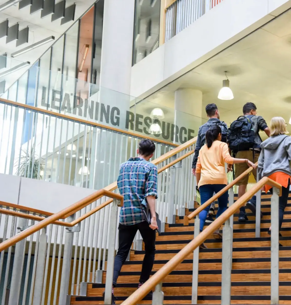Group of students walking up wooden stairs inside a modern building with a glass wall labeled 'Learning Resource'.