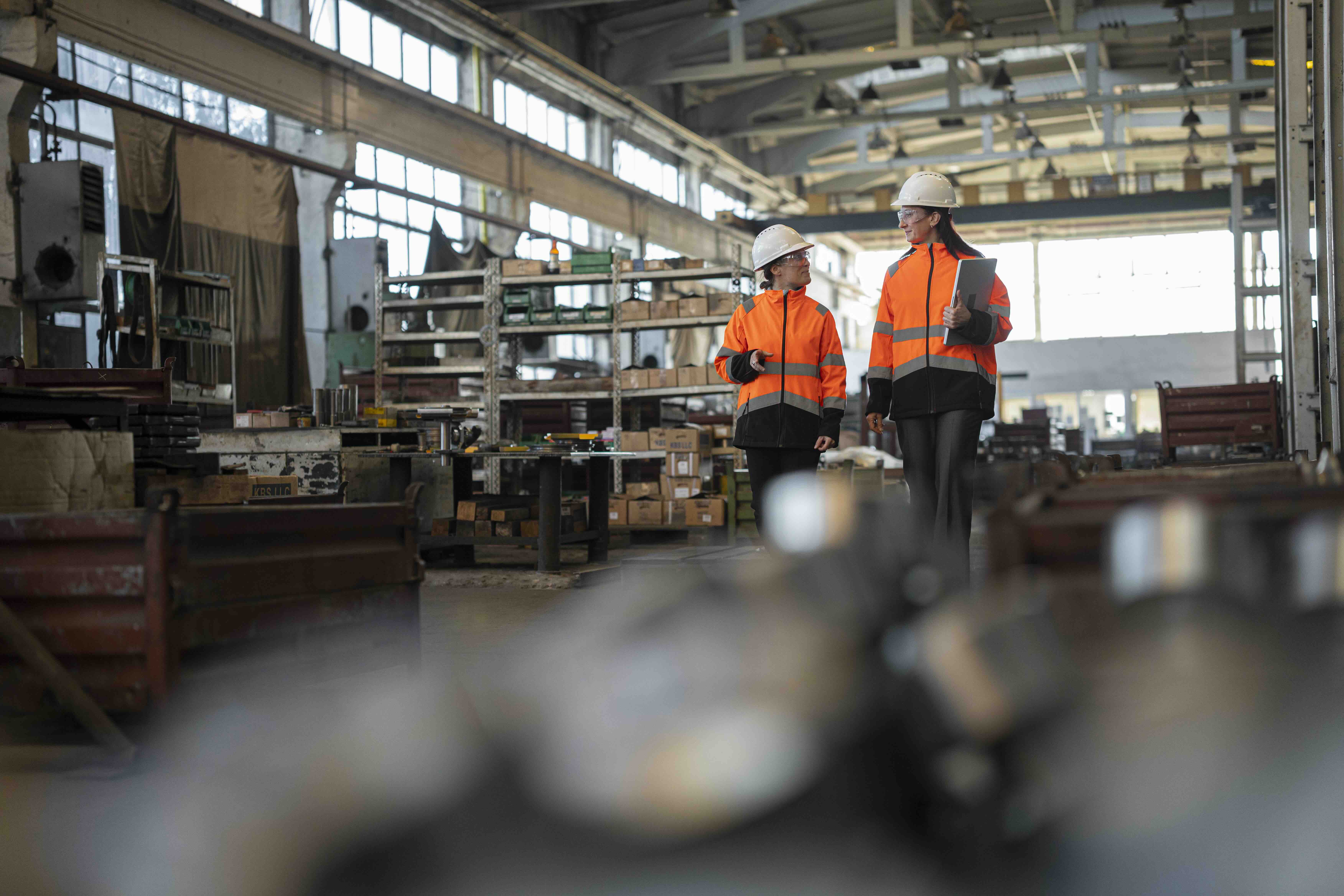 Two factory workers in orange safety jackets and white helmets walking and talking inside an industrial warehouse.