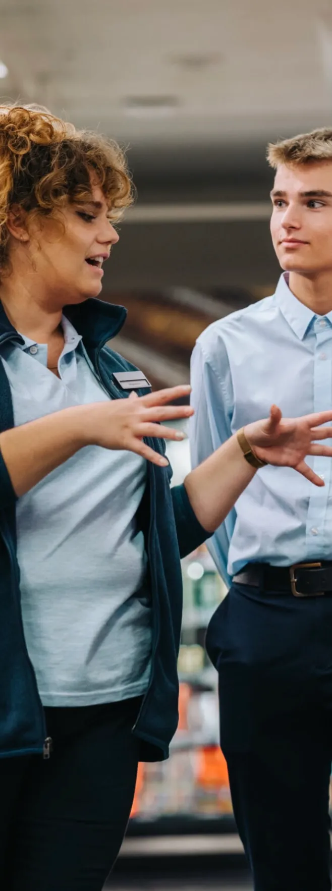 A woman with curly hair and a name tag is explaining something to a young man wearing a light blue shirt and dark pants in a retail setting.