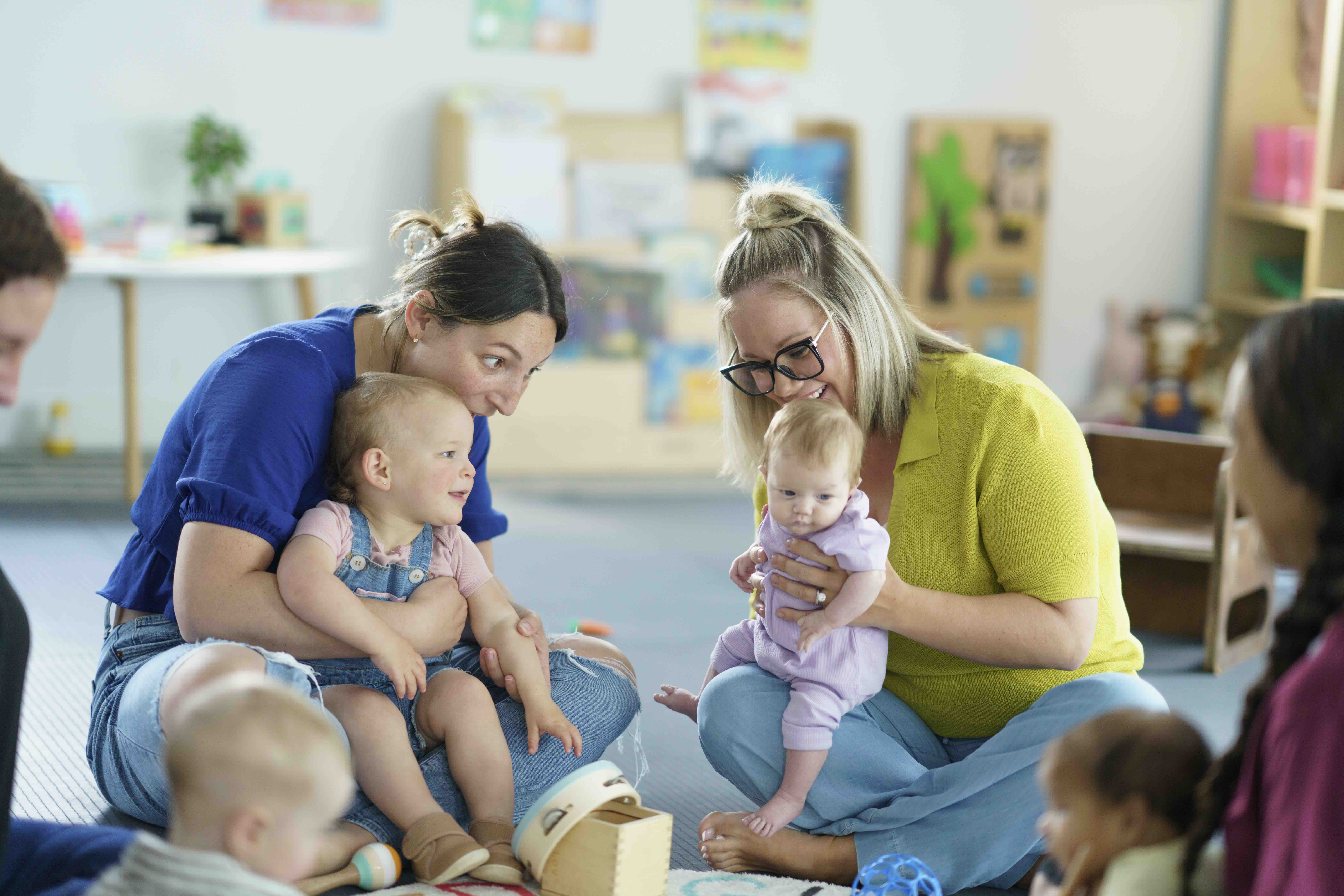 Two women sitting on the floor engaging with babies in a playroom filled with toys and child-friendly decor.