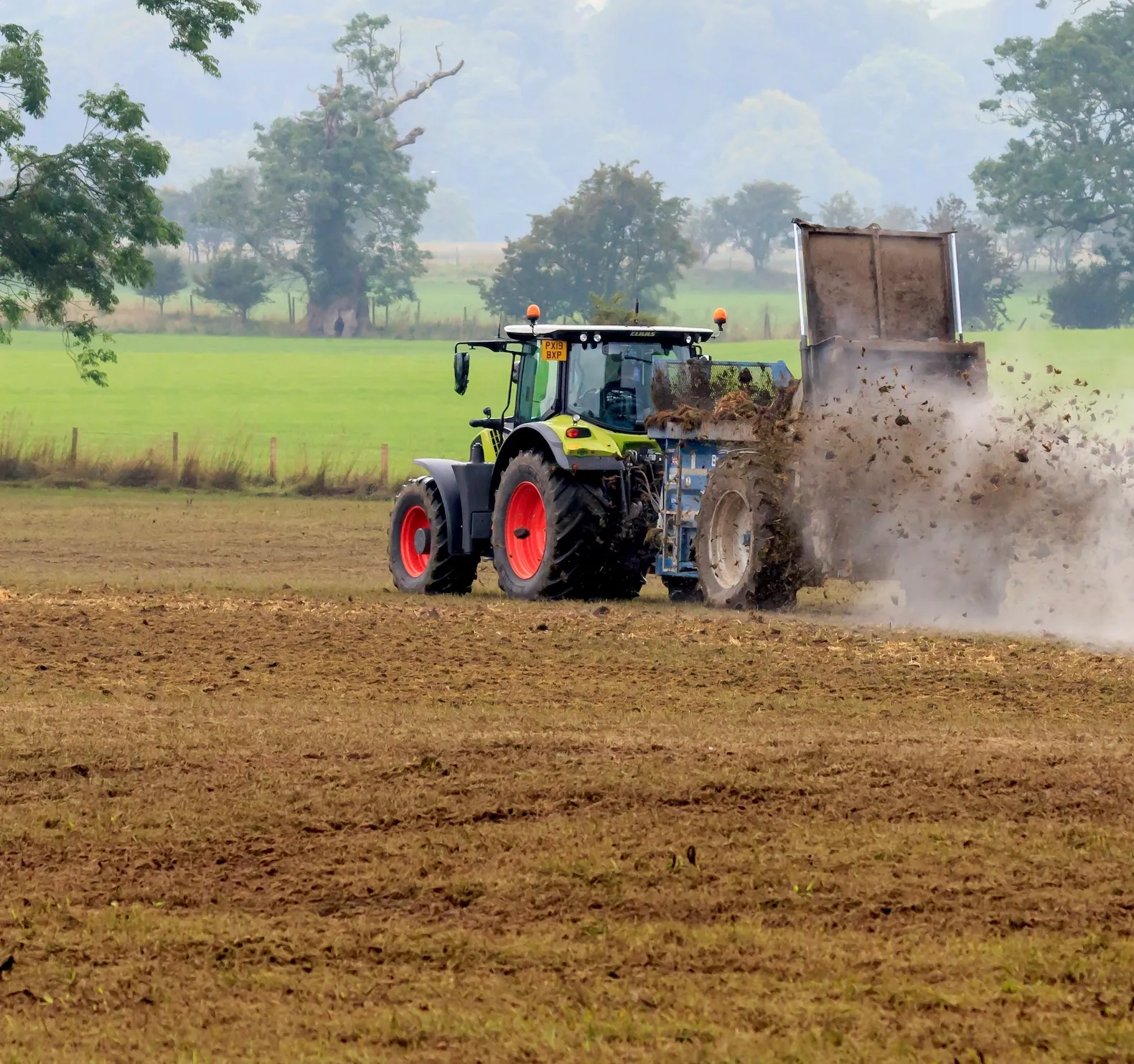 Tractor spreading manure on a plowed field with green trees and grass in the background.