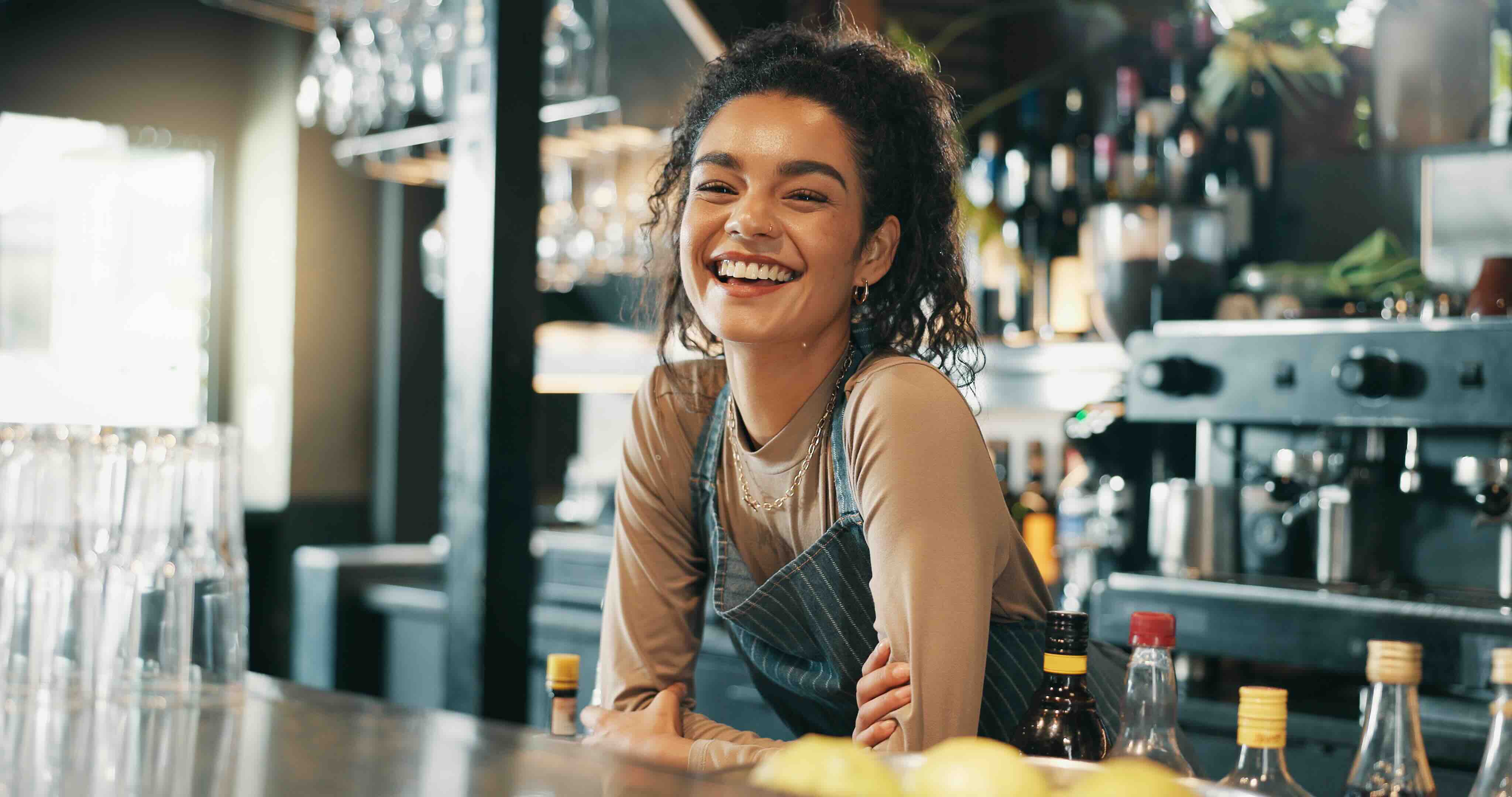 Smiling young woman bartender leaning on a bar counter with bottles and glasses in the background.