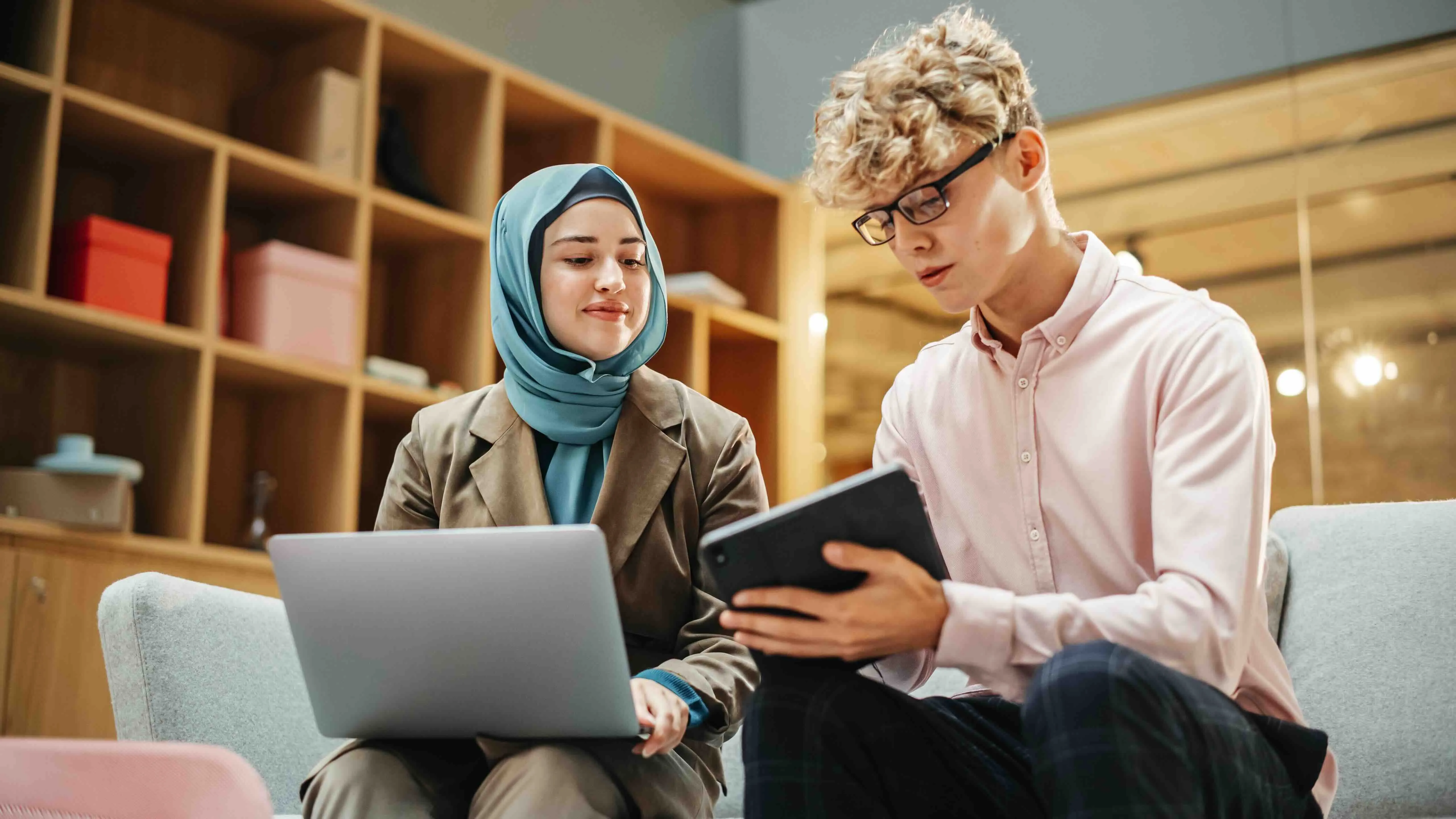 Young woman wearing a hijab and a blazer working on a laptop, sitting next to a young man with glasses holding a tablet in a modern office.
