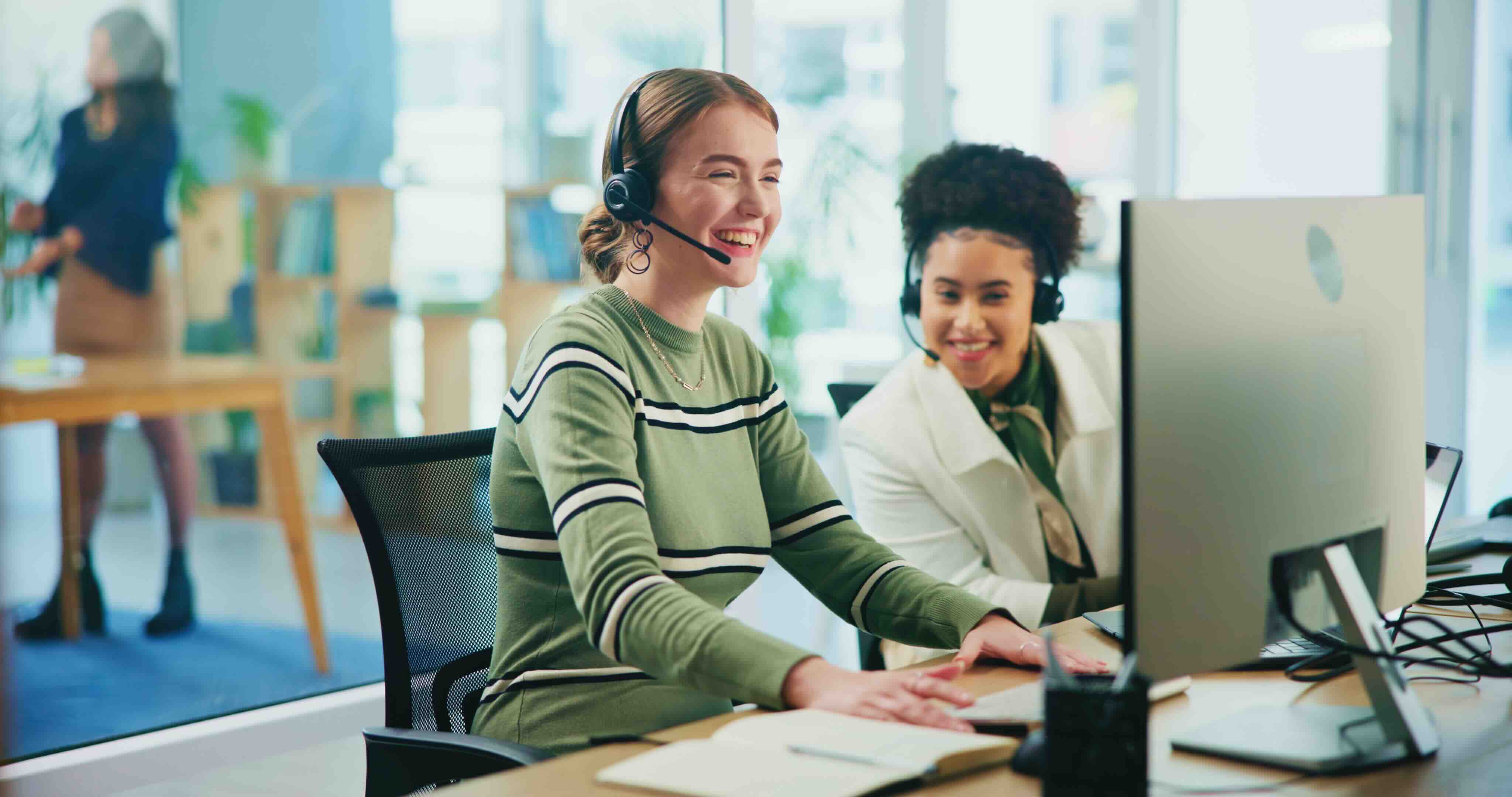 Two women wearing headsets smiling and working at a computer in a bright modern office.