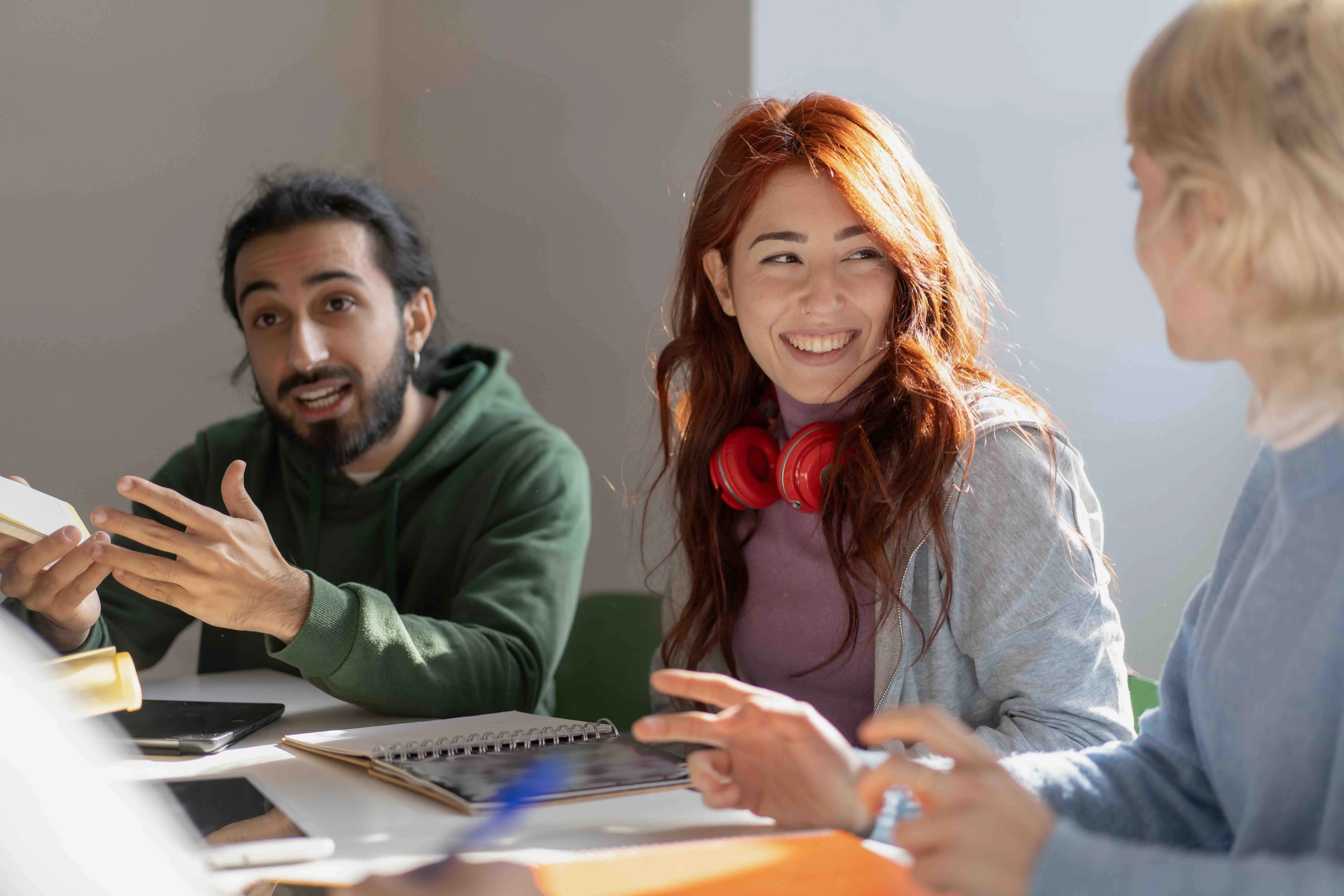 Three young adults engaged in a lively discussion around a table with notebooks and electronic devices.