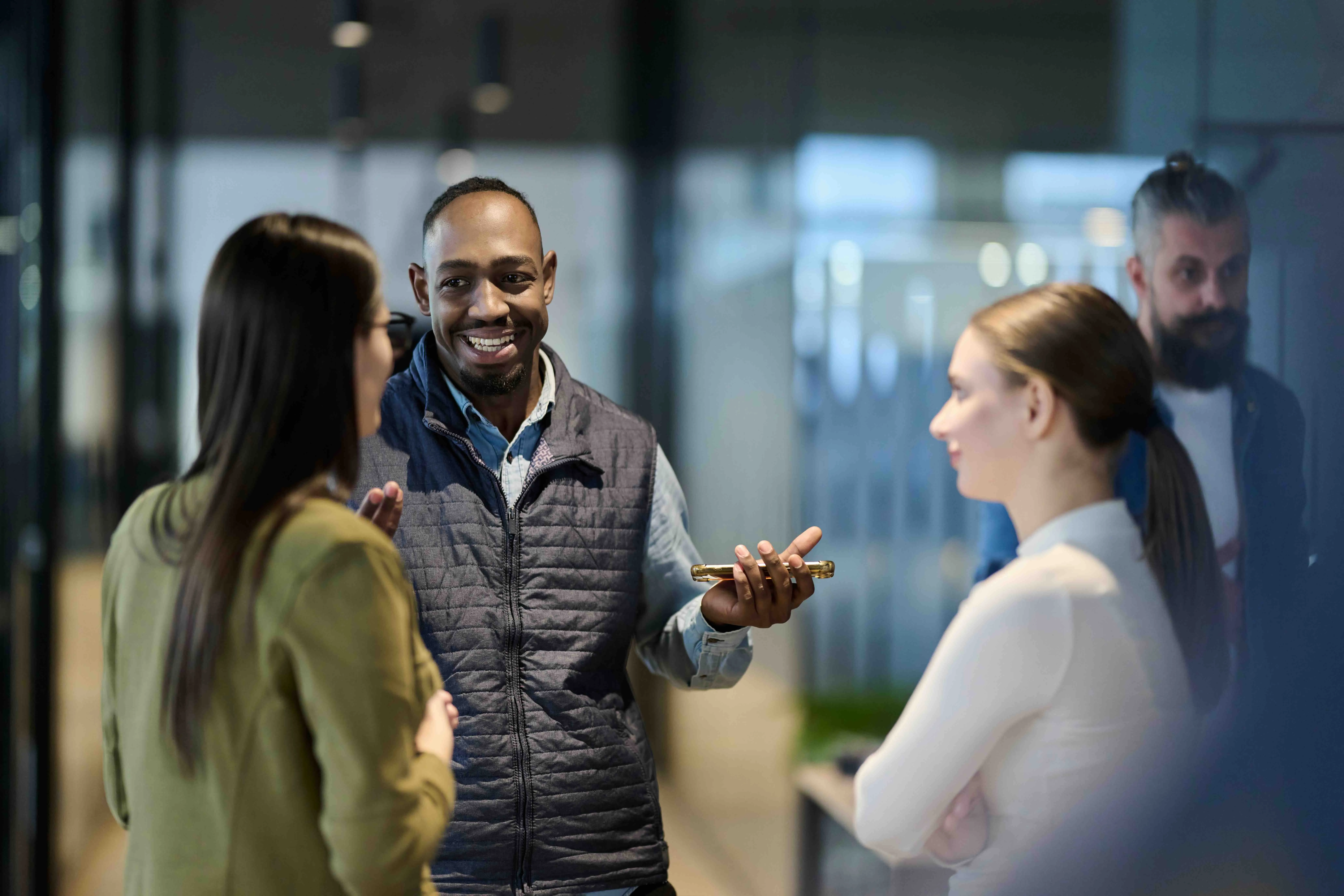 Smiling man holding a phone talks with two women while another man observes in a modern office setting.