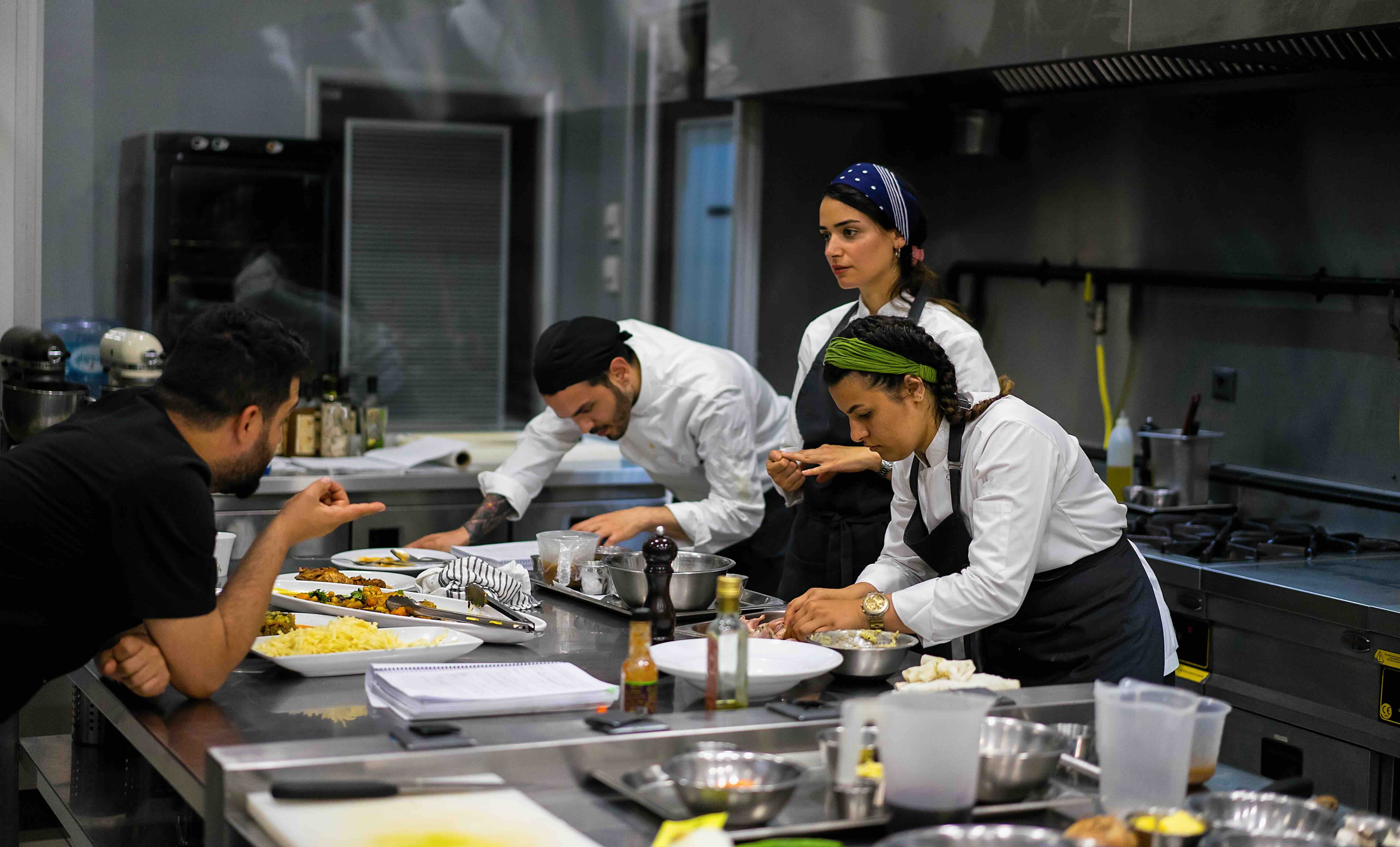 Three chefs and a man in a professional kitchen preparing and discussing plated dishes on a stainless steel counter.