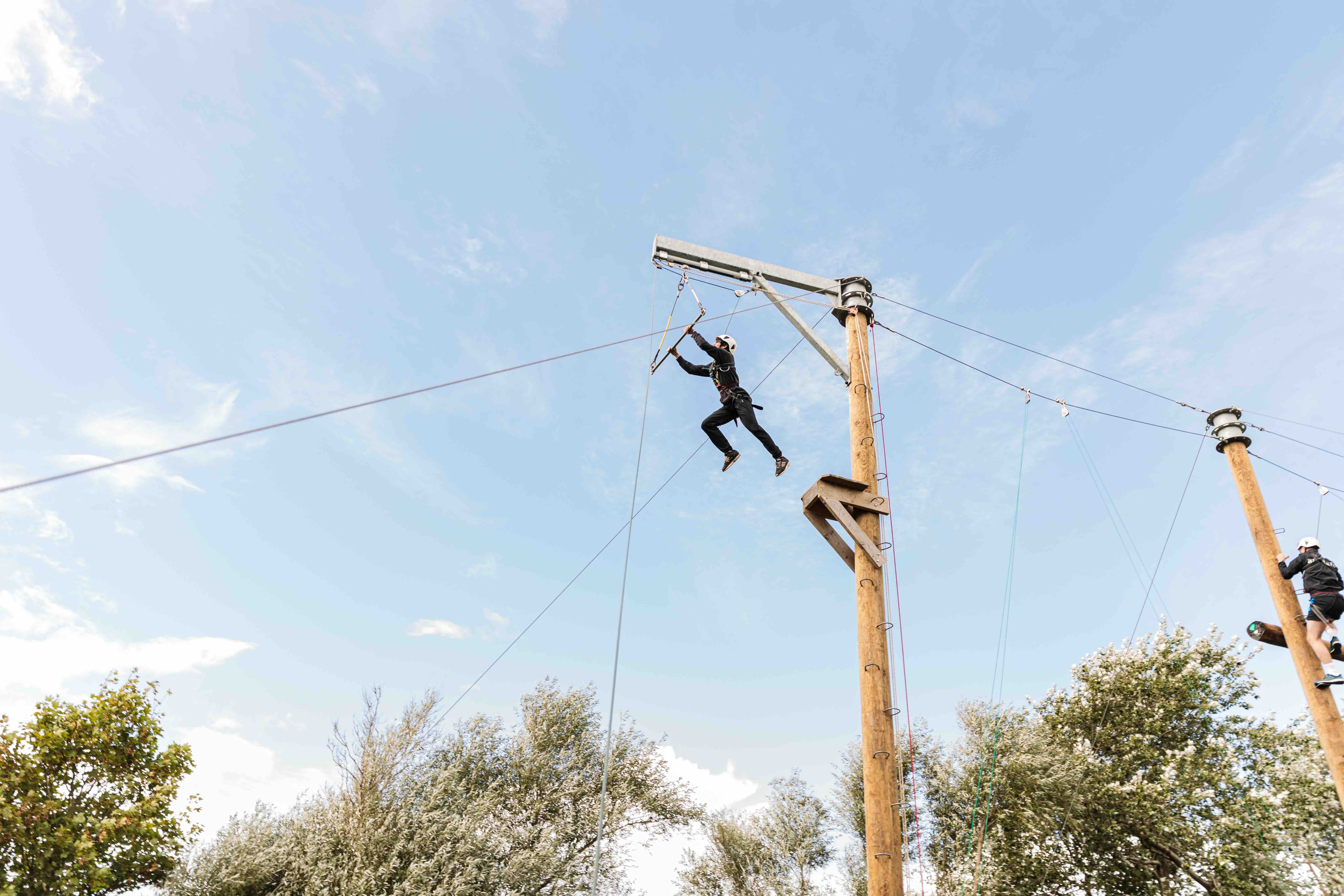 Person wearing safety gear jumping from a wooden platform on a ropes course against a blue sky.