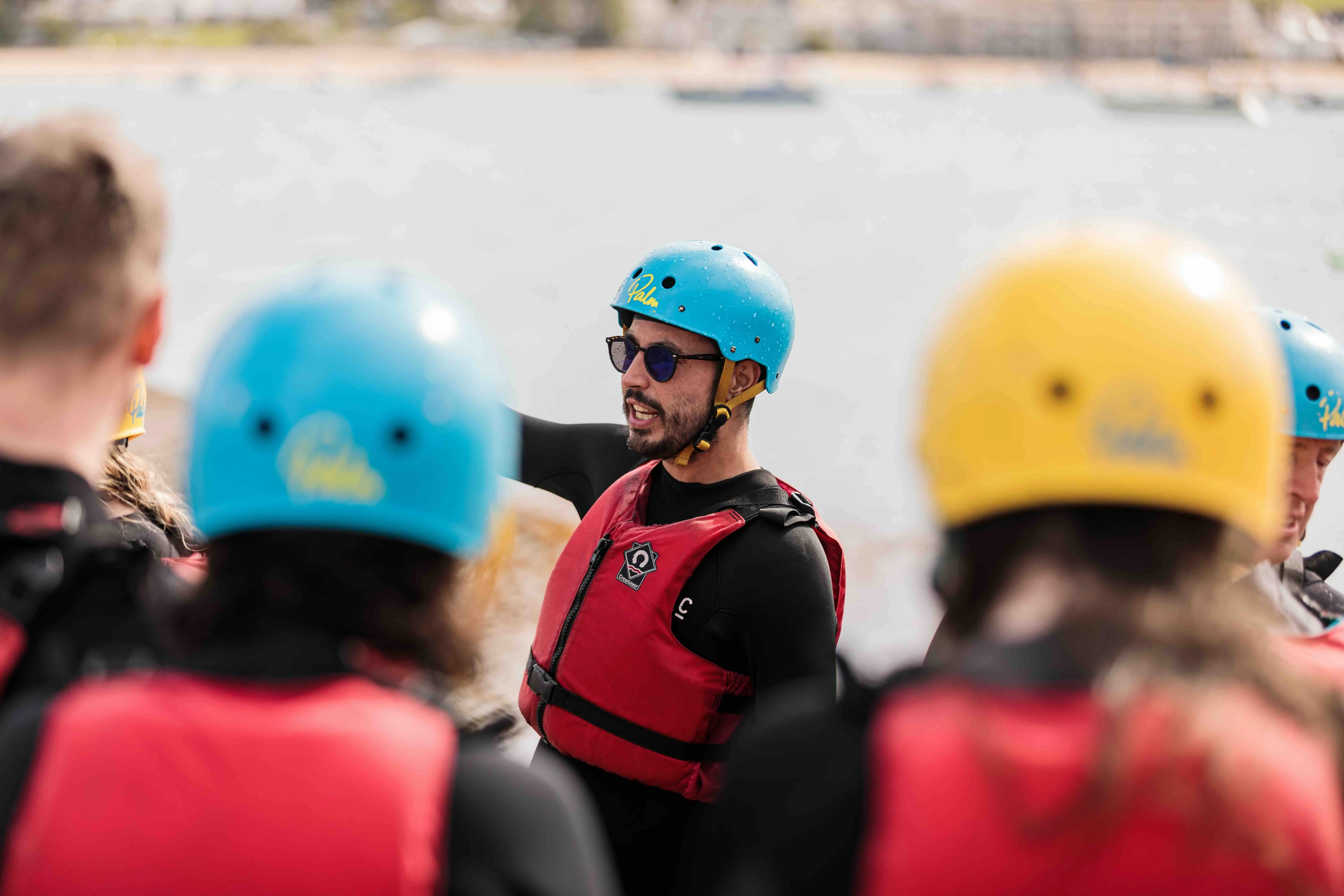 Man wearing blue helmet and red life jacket giving instructions to a group near water.