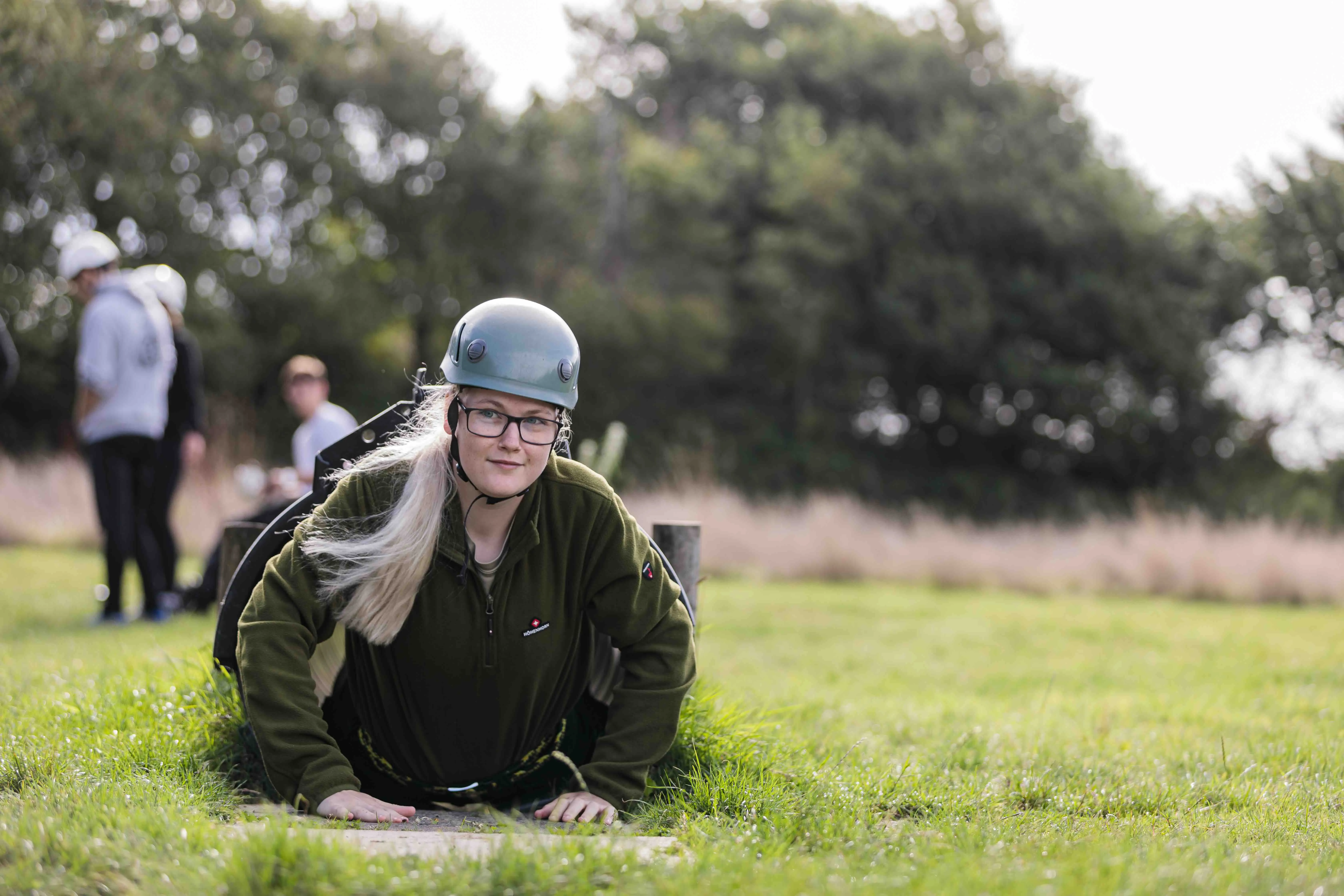 Person wearing a helmet and glasses crawling out of a small tunnel on grass in an outdoor setting.