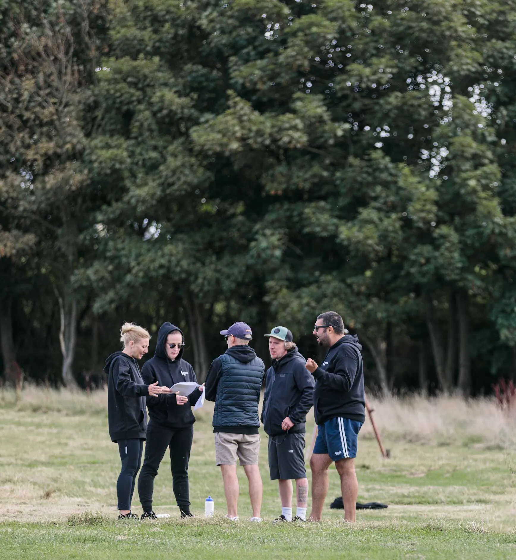 Group of five people standing on grass outdoors with trees in the background, some holding papers and discussing.